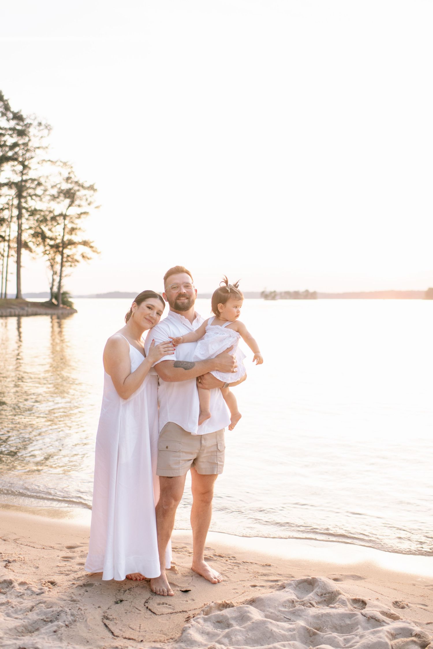 Family embracing on beach with cypress trees and sunset water in background.