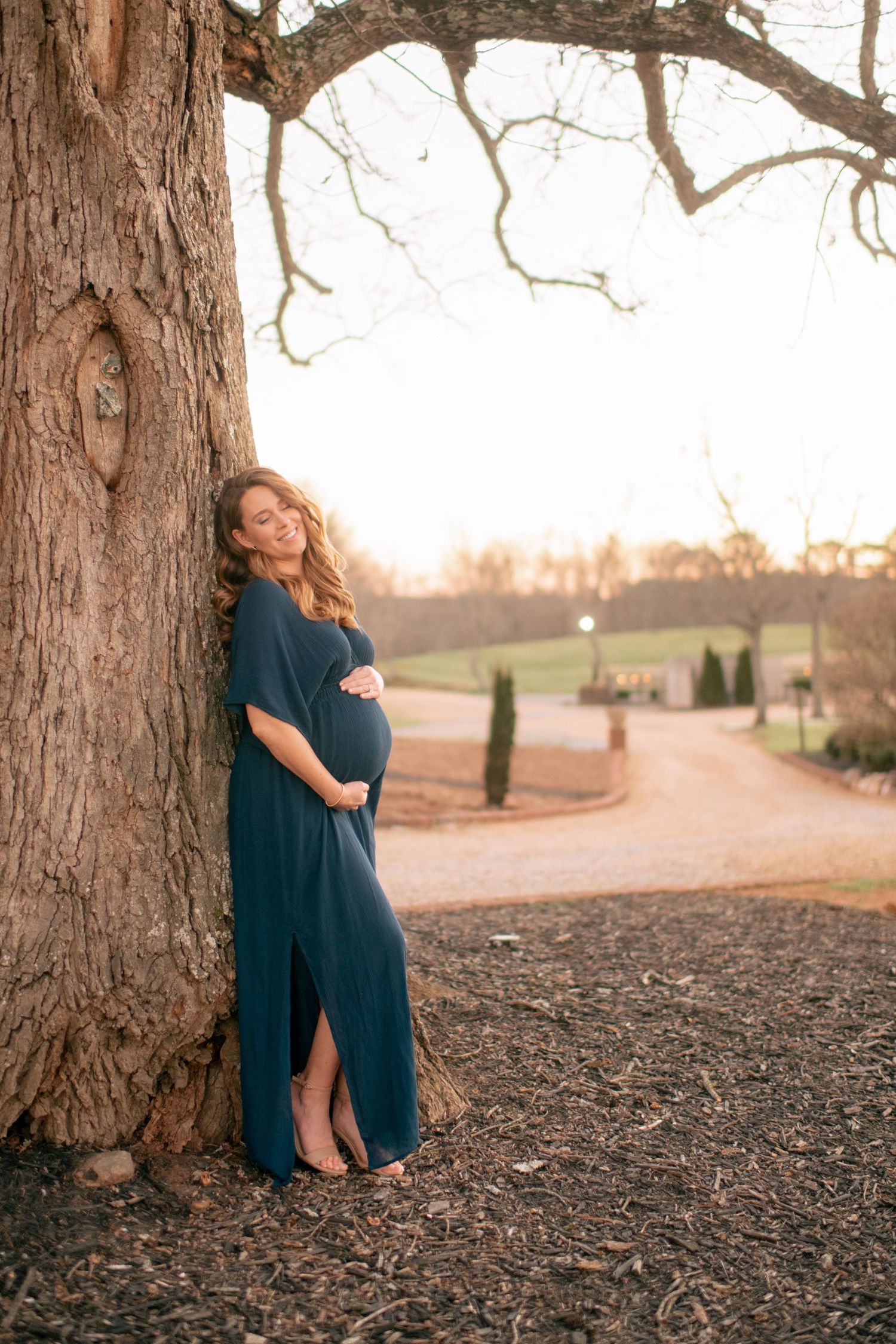 Expectant mother in flowing navy dress poses against large tree trunk during golden hour sunset in outdoor setting.