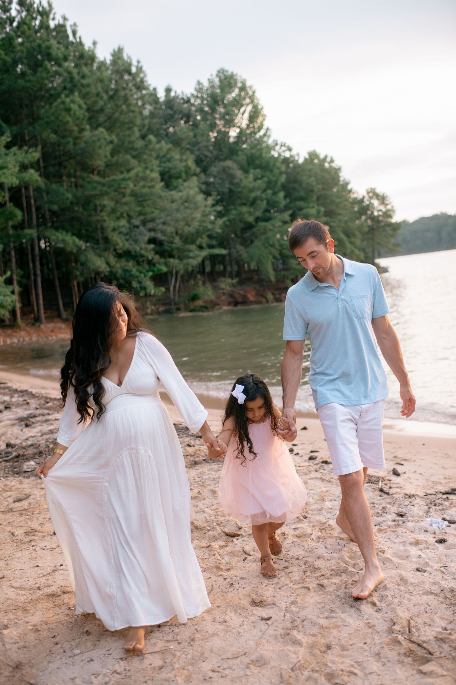 A family walks barefoot together along a sandy beach by a lake surrounded by pine trees on a cloudy day.