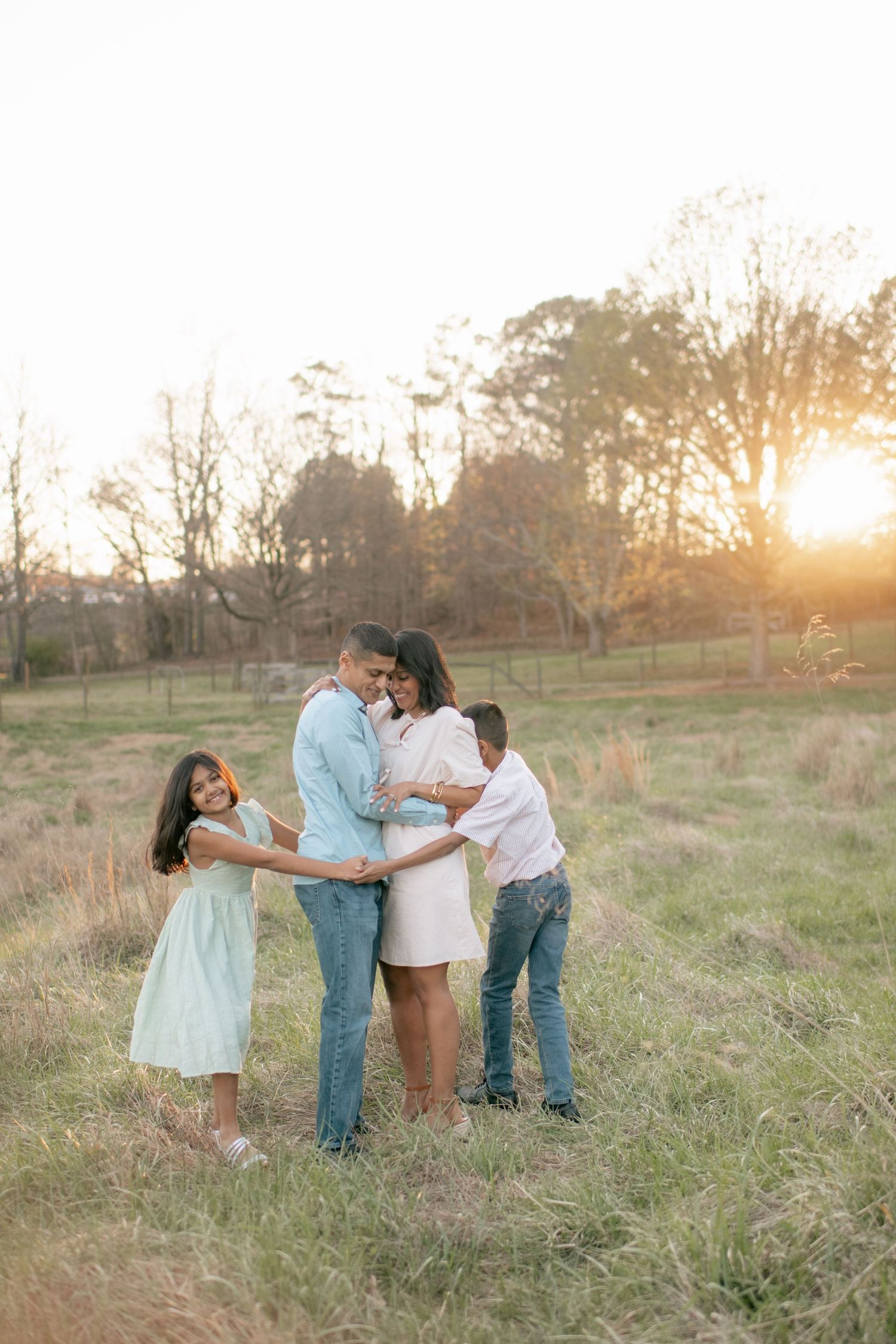 Family of four embraces in a golden field during sunset with trees in background.