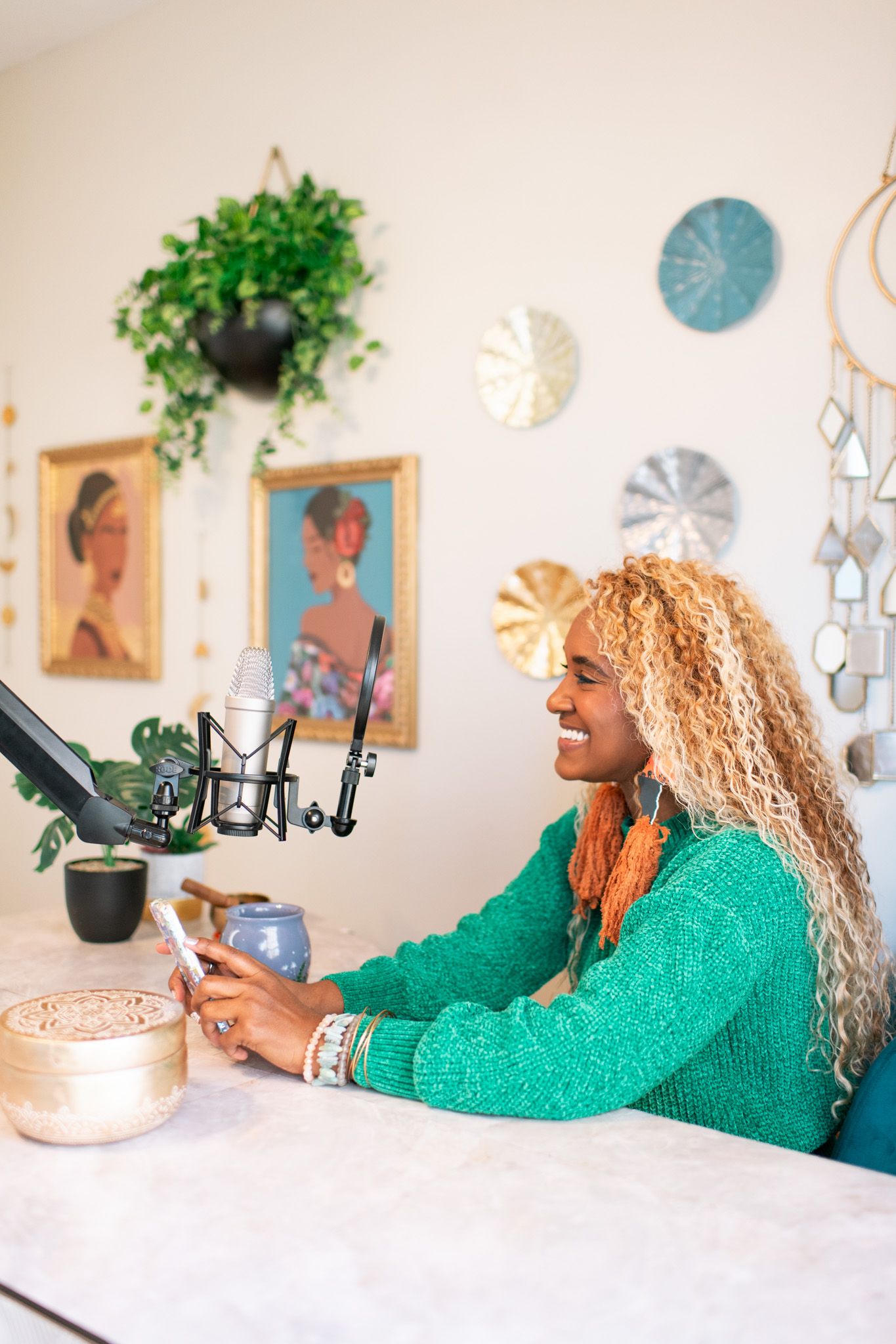 Smiling woman with blonde curly hair in green sweater podcasting at microphone with phone in boho studio.