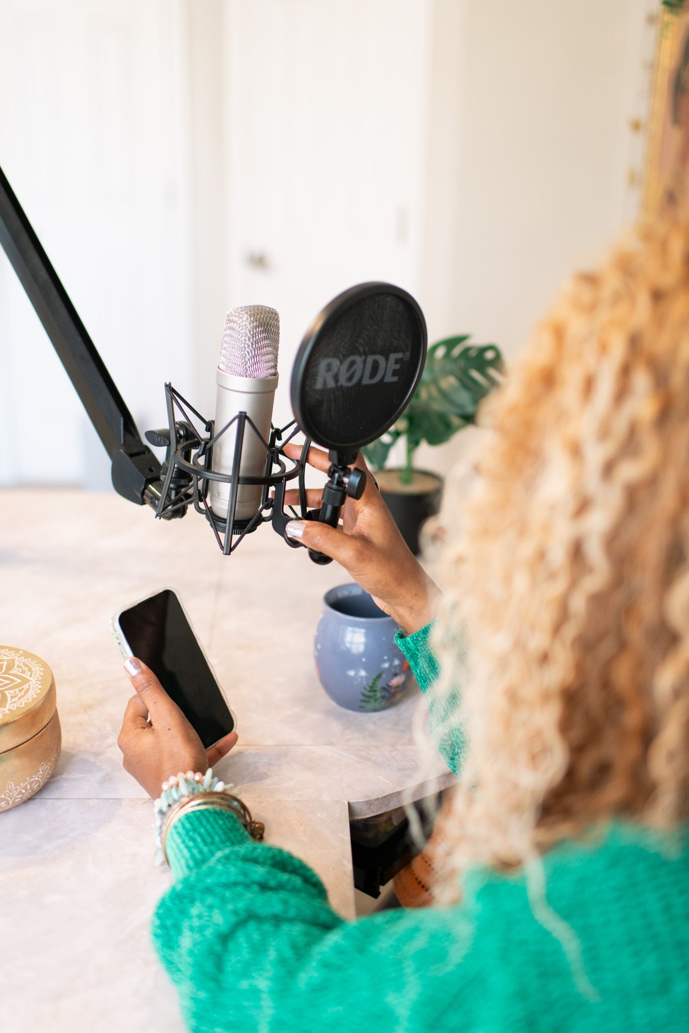 Woman in green sweater holding a RODE microphone and smartphone at a home recording studio setup.