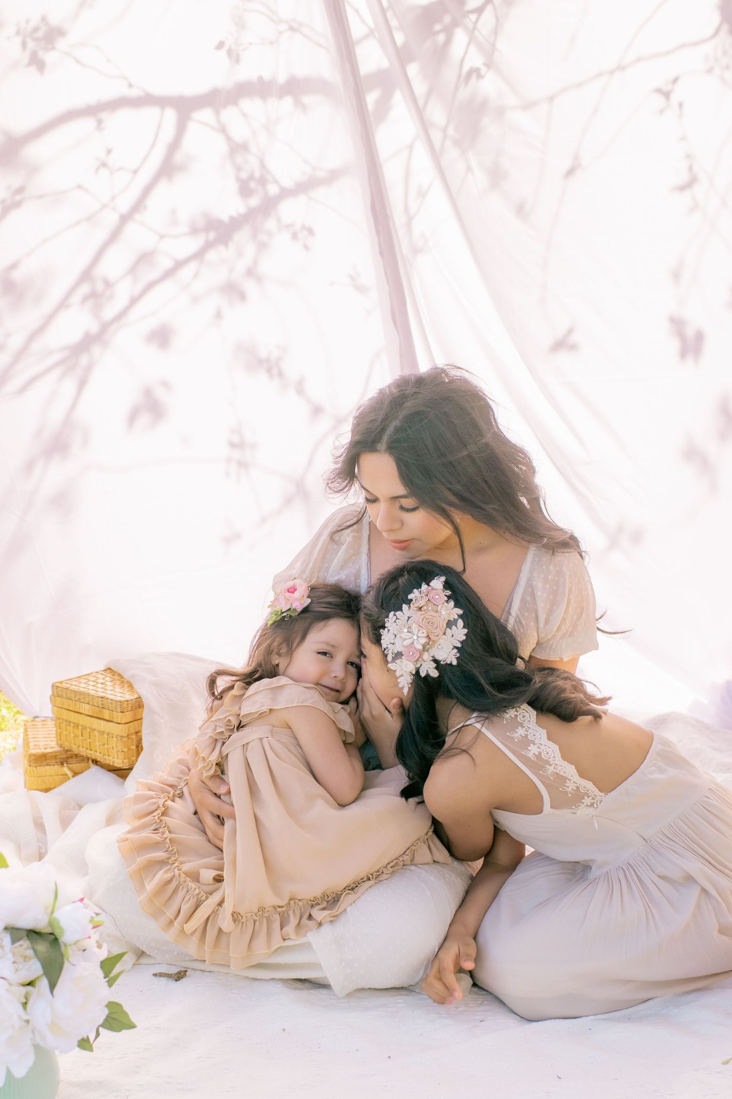 A dreamy mother-daughter portrait with soft pink tones shows them cuddling on white cushions with spring flowers and a basket nearby.