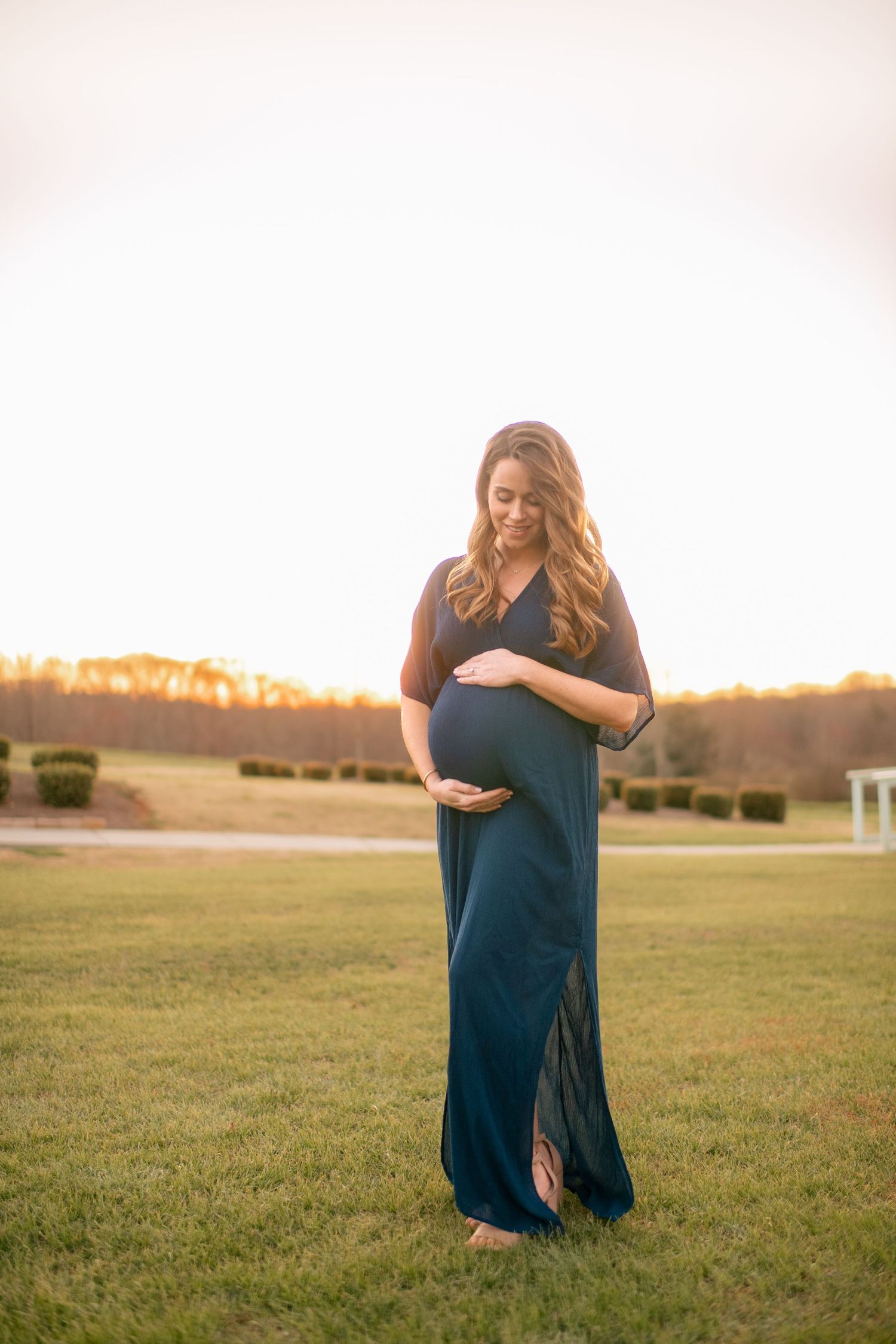 An expectant woman in a navy maxi dress stands barefoot in a grassy field at sunset.