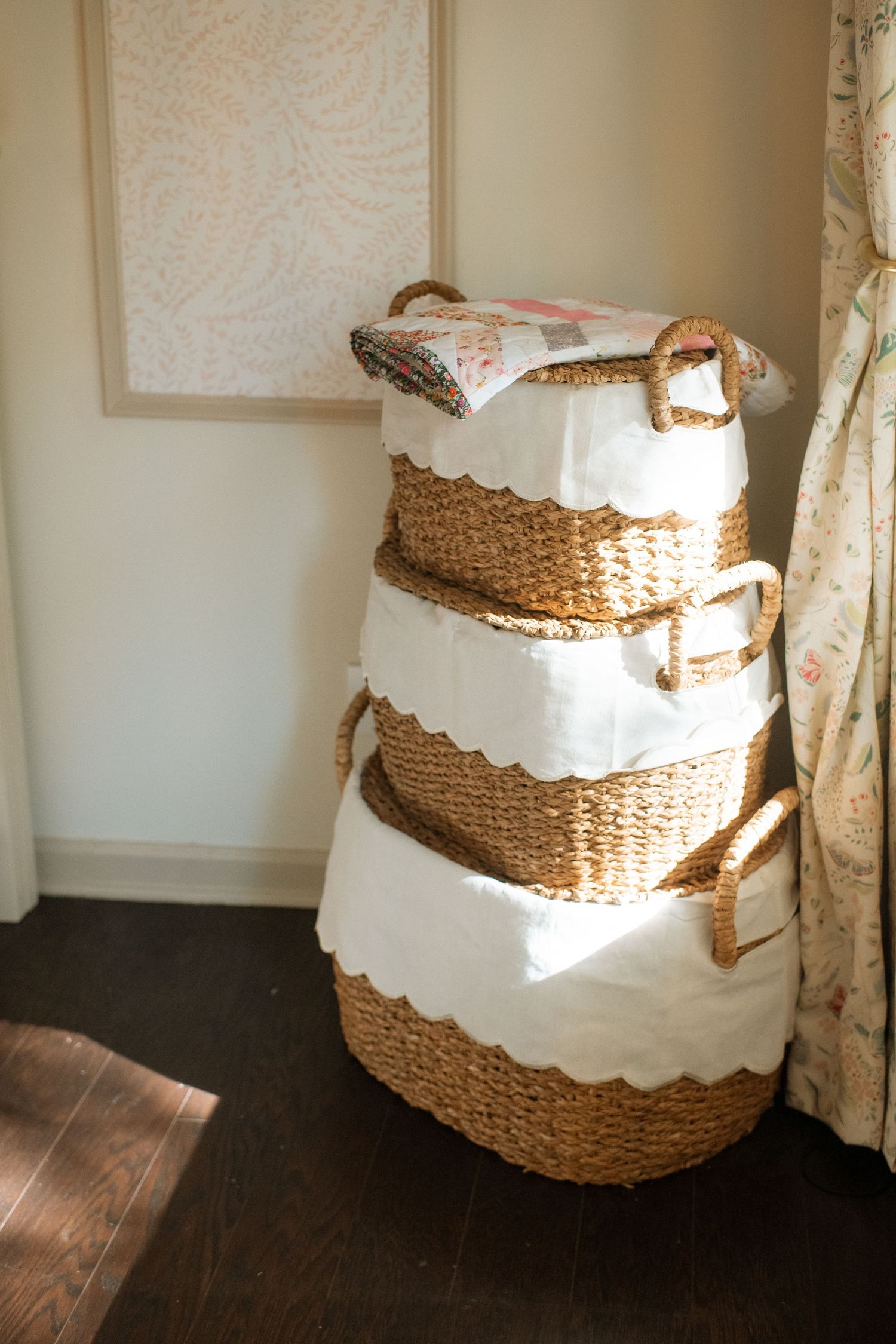 Three stacked woven storage baskets with white trim sit in a corner of a room.