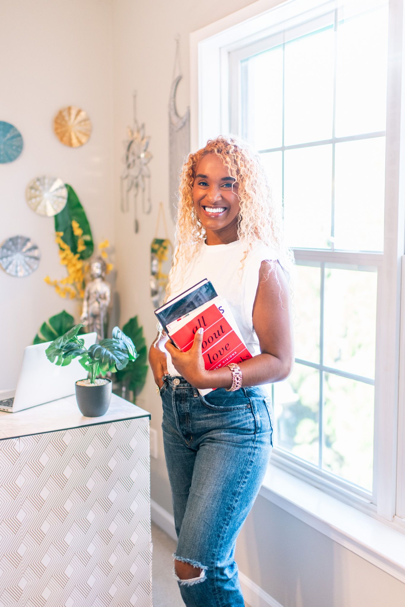 Smiling woman with curly blonde hair holding books, wearing white top and ripped jeans, standing by a bright window indoors.