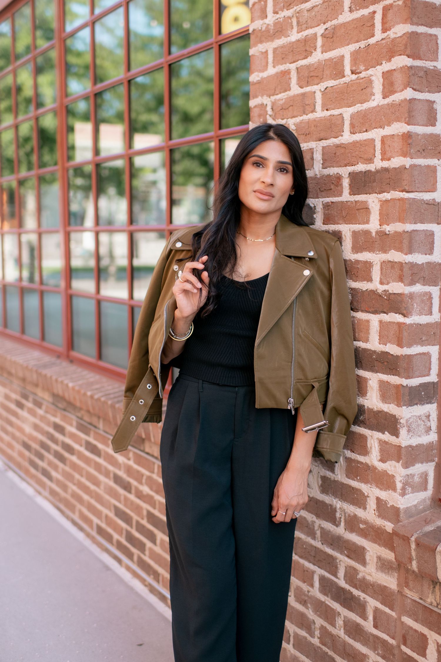 A person poses against a brick wall wearing a black jumpsuit and brown leather jacket in a fashion photoshoot series.