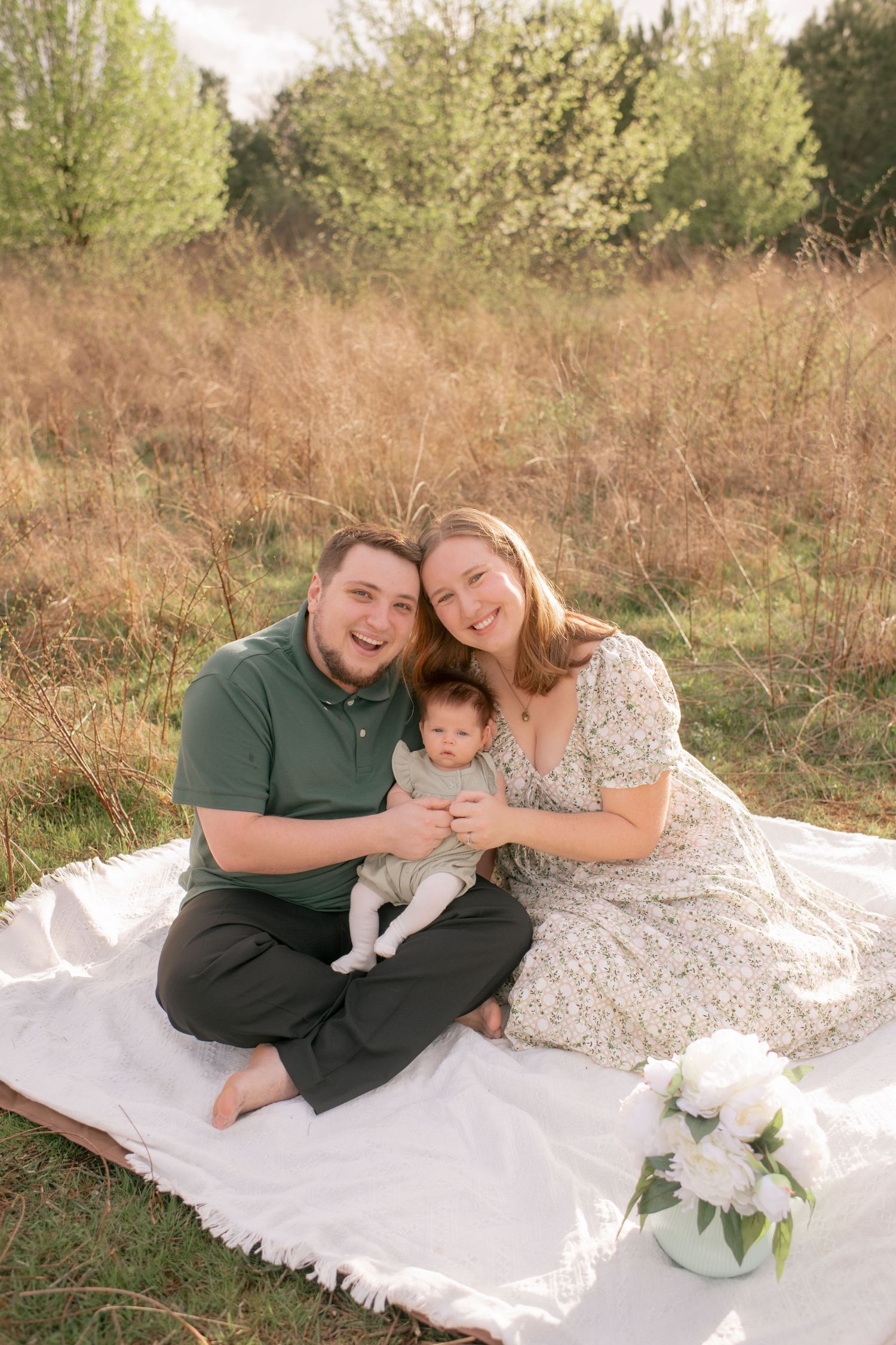 A family sits together on a white blanket in a grassy field during golden hour with flowers in a vase nearby.