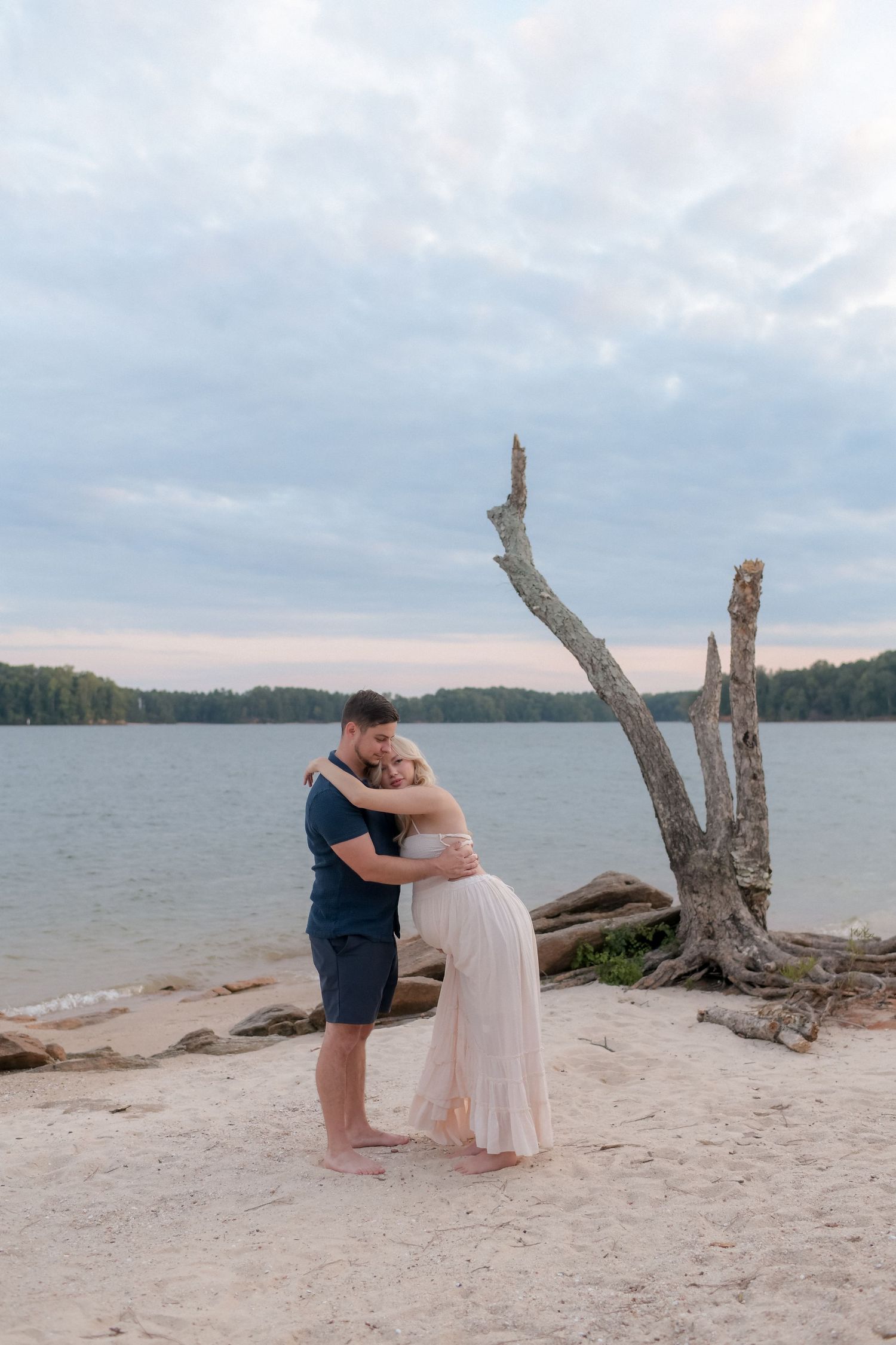 A romantic maternity photoshoot on a sandy beach with driftwood and calm waters.