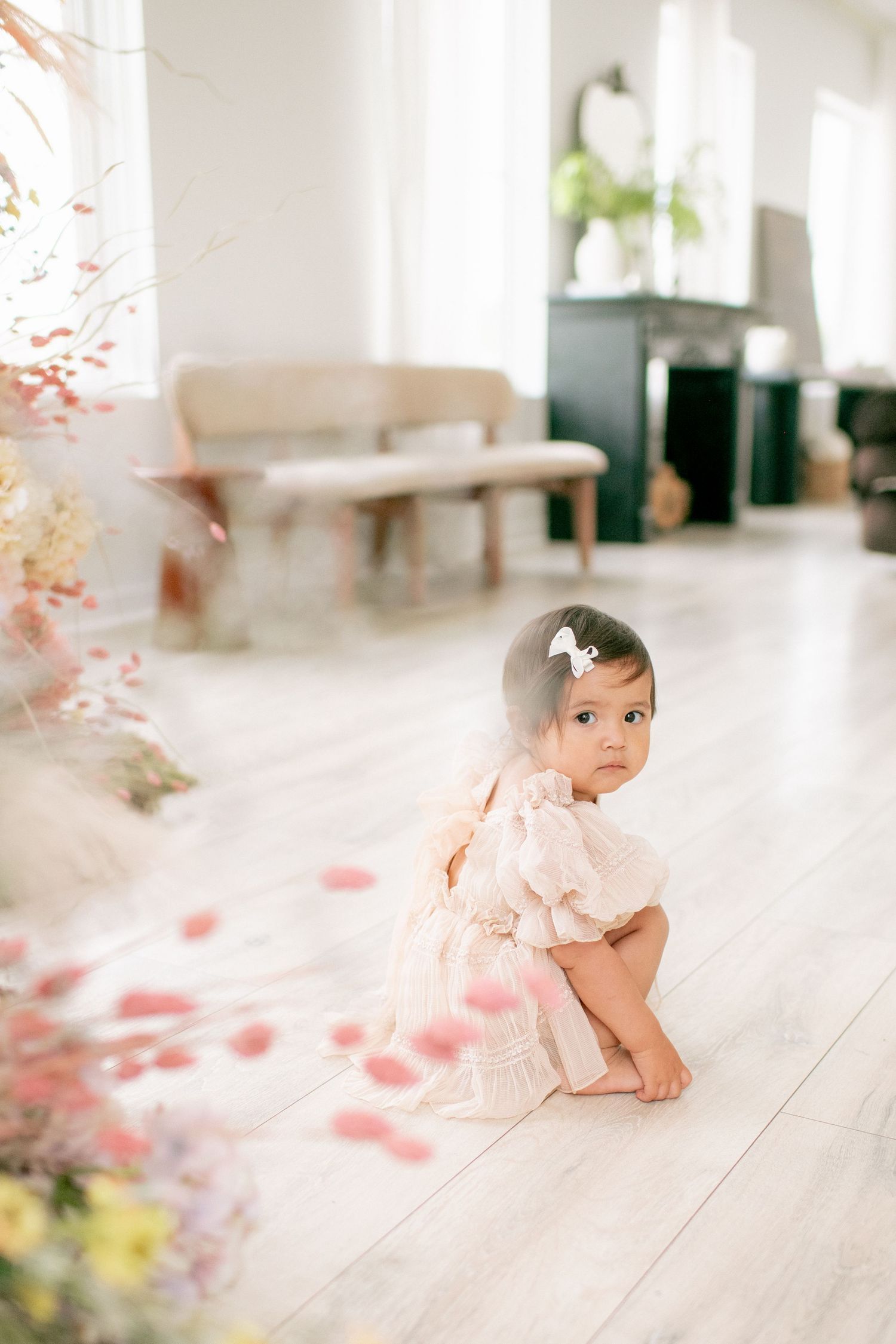 A small child in a cream colored dress crouches on a light wood floor with soft pink floral decor in the foreground.