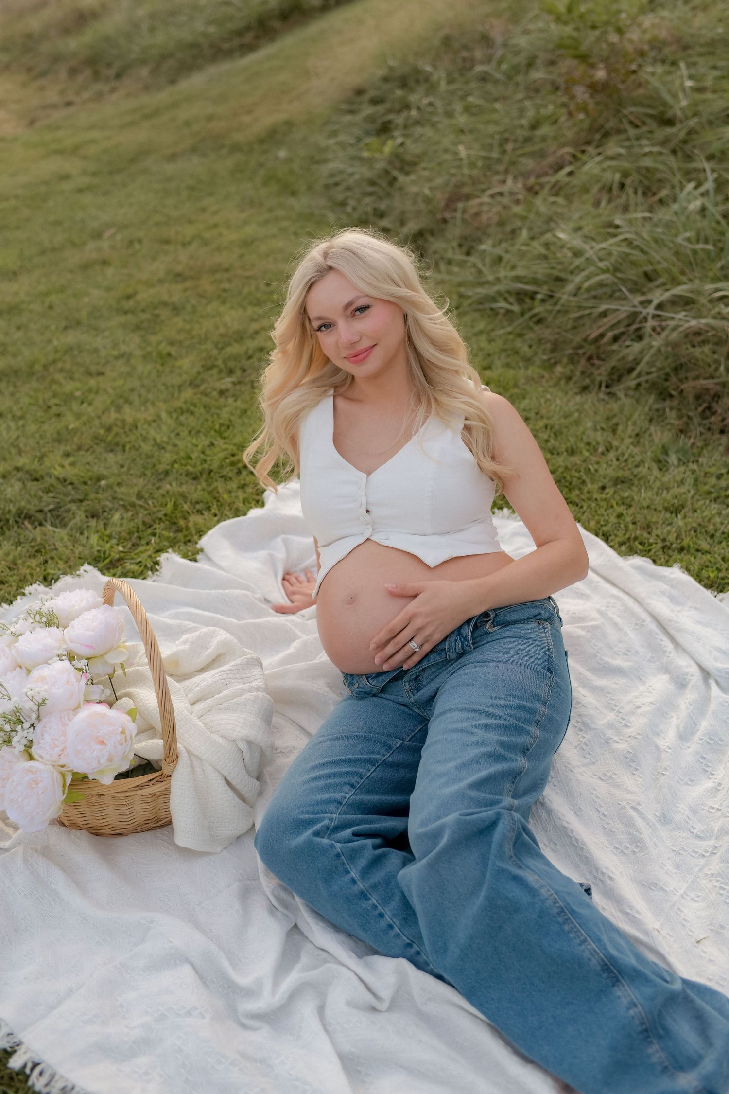 Expectant mother in white top and jeans poses on white blanket during outdoor photoshoot.