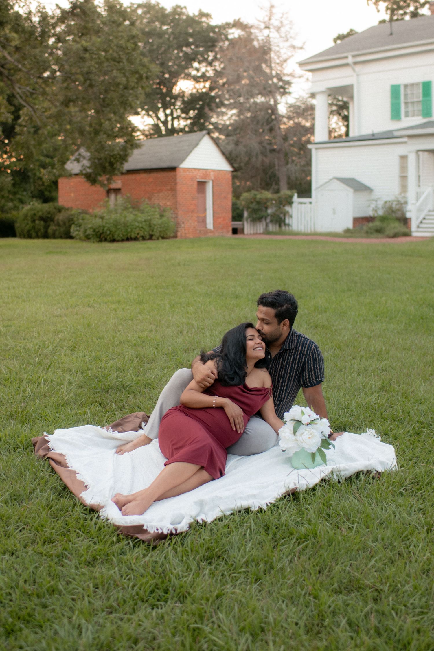 A couple lying on a white blanket in the grass sharing an intimate moment near a white building.