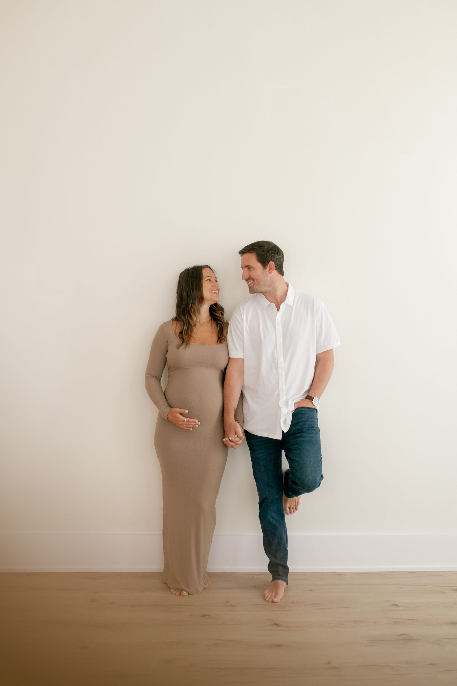 Couple stands against white wall during minimalist maternity photoshoot.