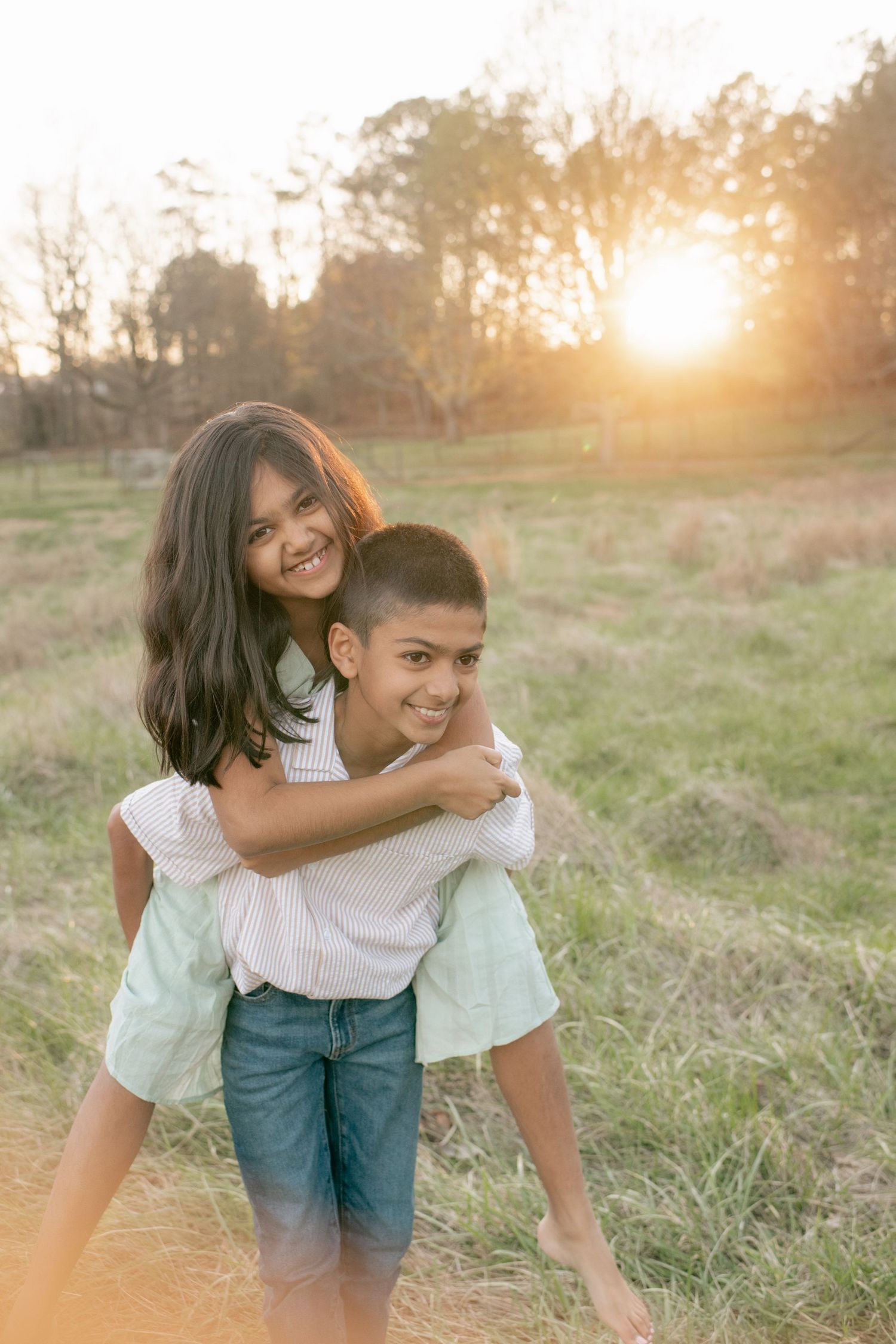 Two siblings share a playful piggyback ride in a sunlit meadow at golden hour.