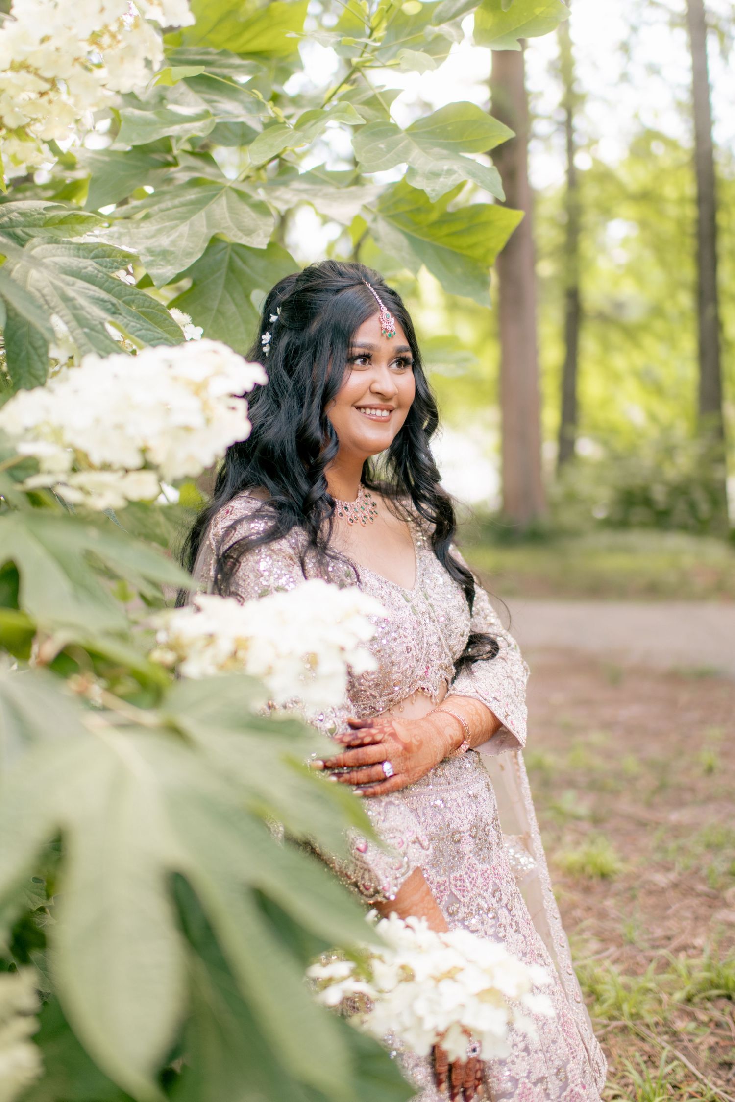 A person in a white lace dress poses gracefully among blooming trees in a sunlit forest setting.