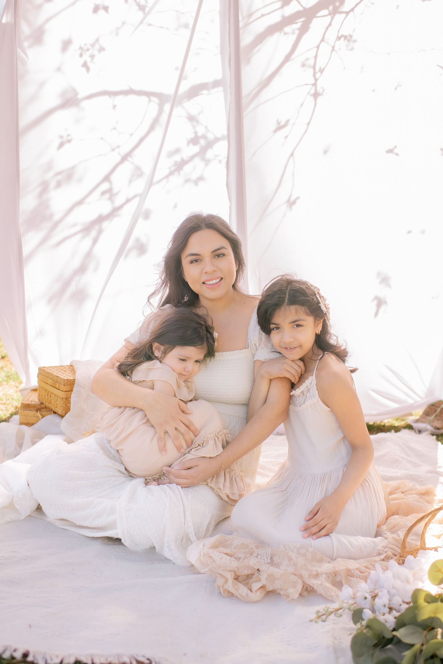 Dreamy springtime family portrait with white dresses and floral accents on a soft blanket in natural lighting.