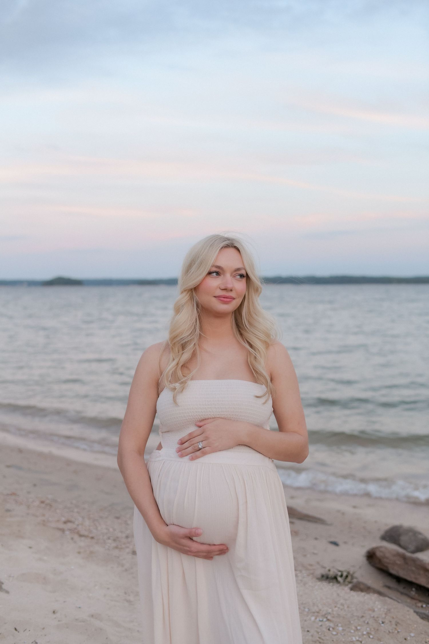An expectant mother poses on a beach in a light dress during golden hour.