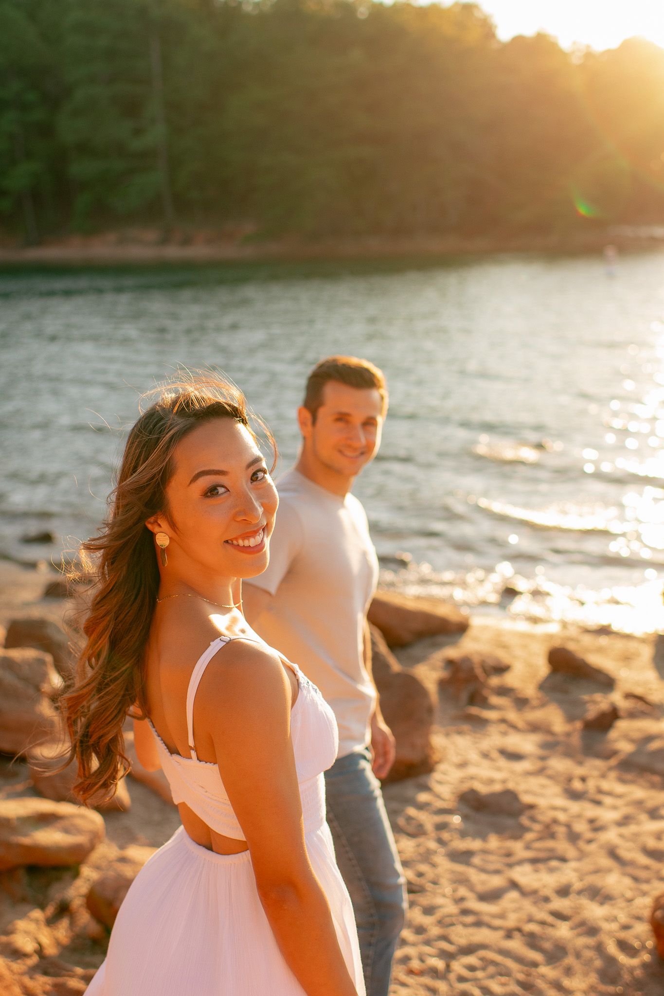 Couple enjoying a romantic sunset moment on a rocky beach shore with golden sunlight and water in background.