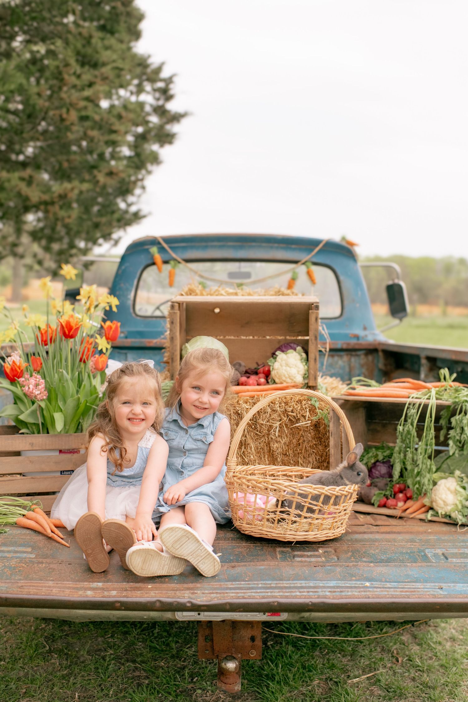 Two children sit on vintage truck bed with spring flowers, hay bales and rustic baskets in country setting.