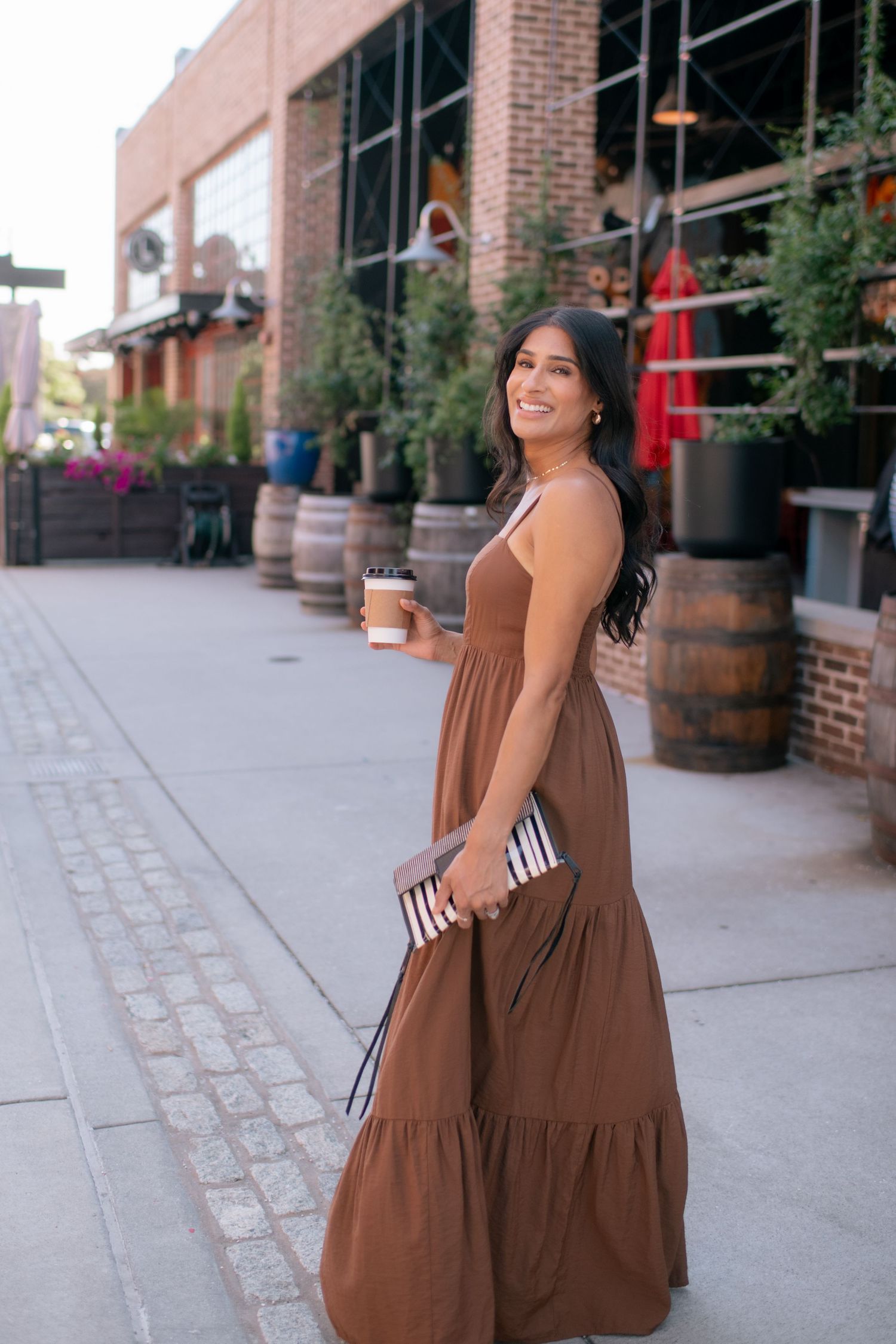 Person in brown tiered maxi dress walking on cobblestone street near brick building with decorative barrels.