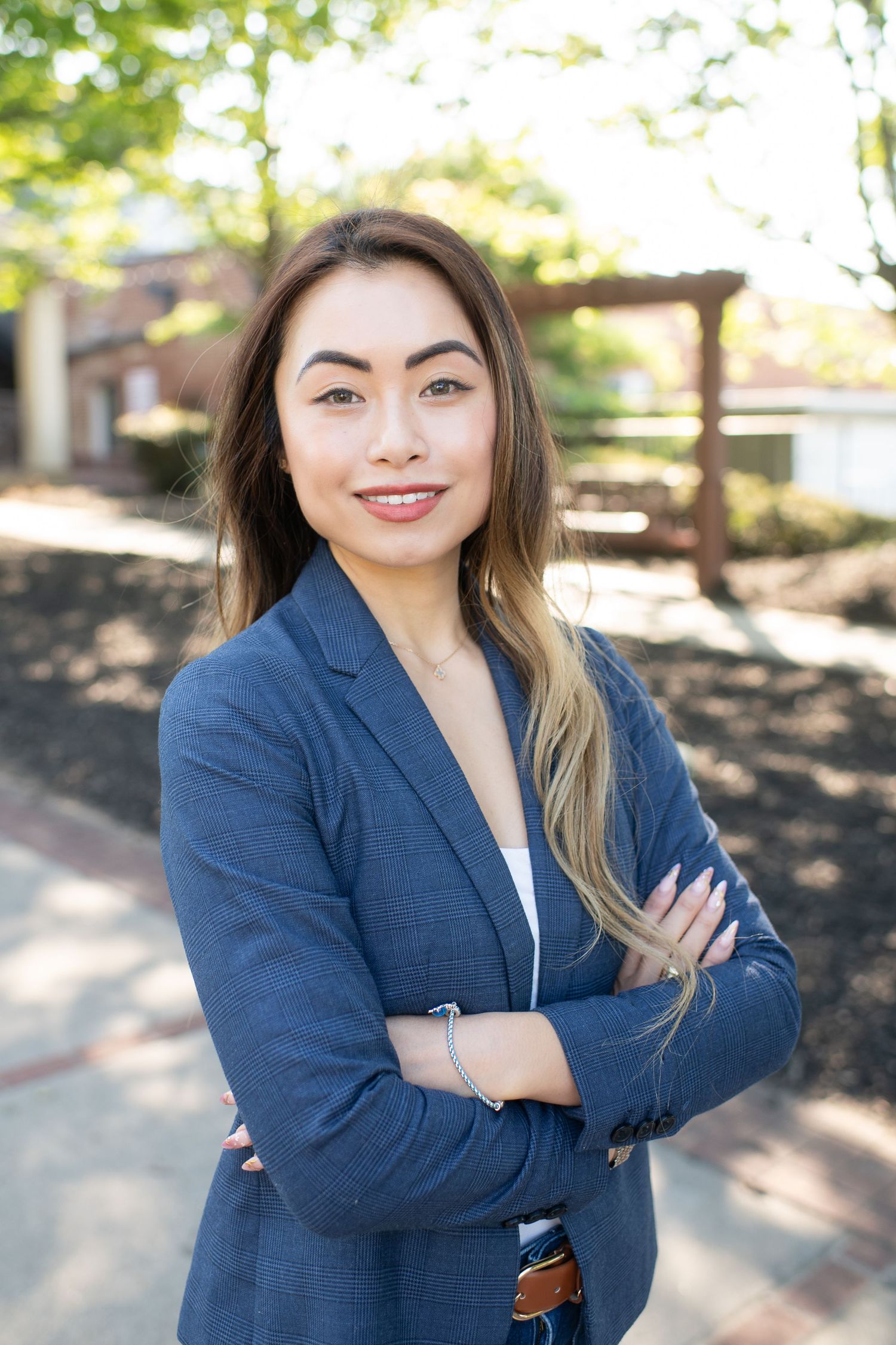 Young professional woman in blue blazer smiling with arms crossed outdoors in a sunny campus setting.