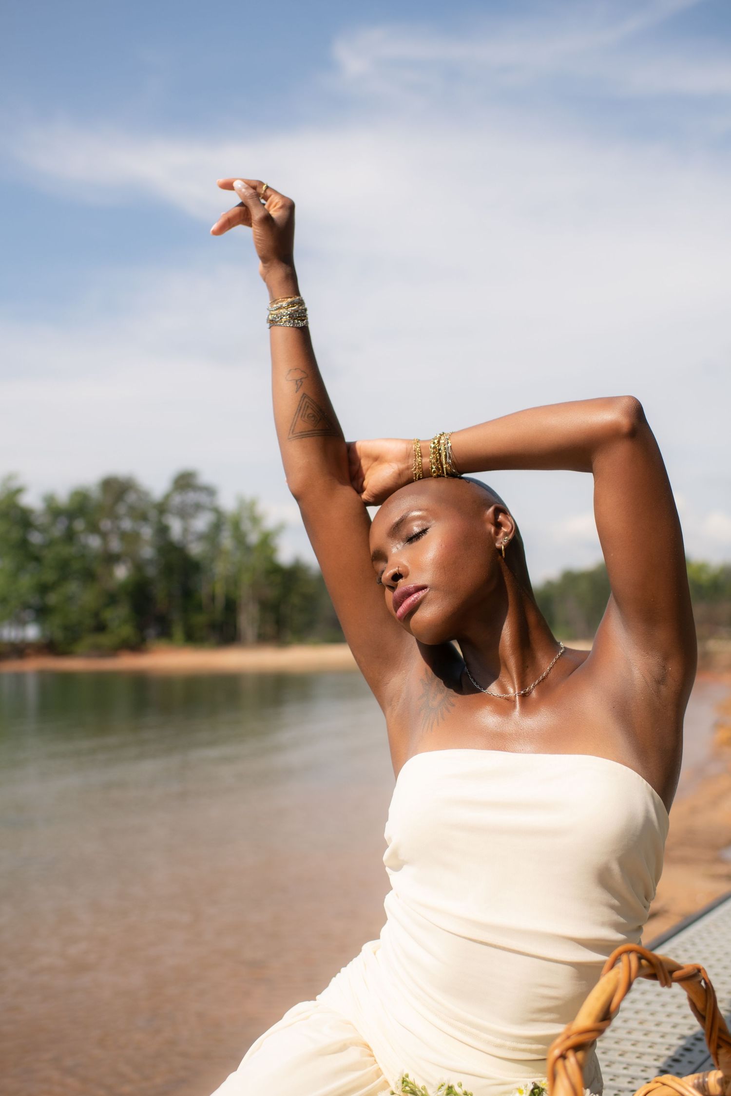 Elegant Black woman in white strapless dress poses gracefully by a lake, arms raised, wearing gold jewelry.