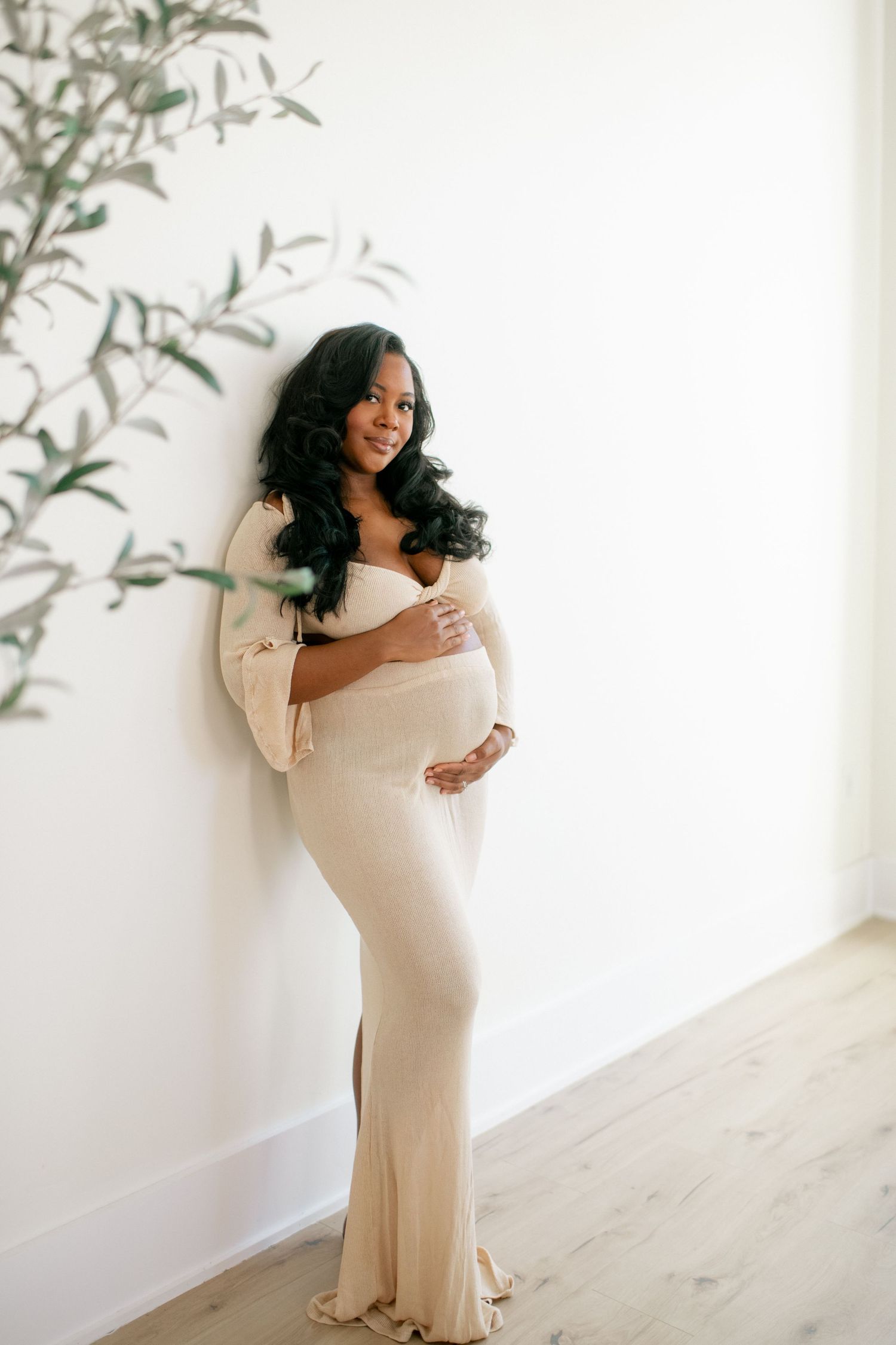 Maternity photoshoot featuring a cream-colored dress against a white backdrop with delicate foliage accents.