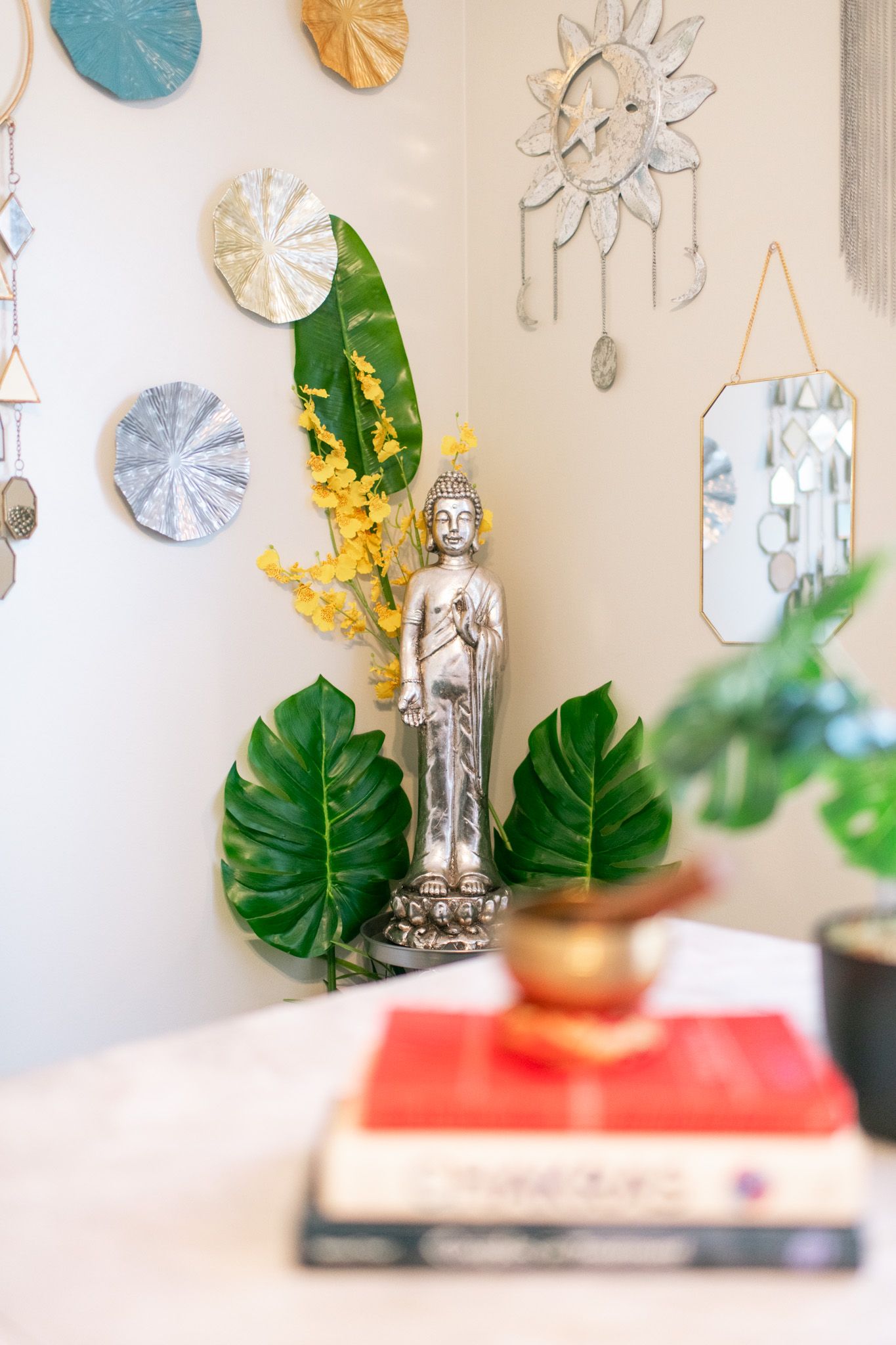 Decorative wall display featuring metallic sun shapes and green monstera leaves with small Buddha statue.