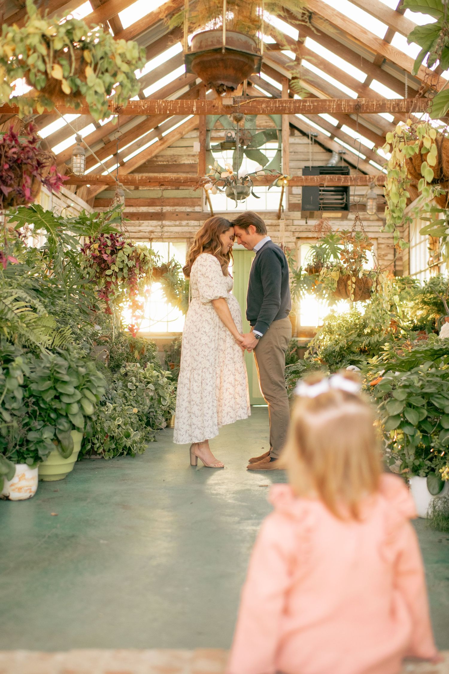 A romantic moment captured in a sunlit greenhouse filled with lush plants and hanging flowers.