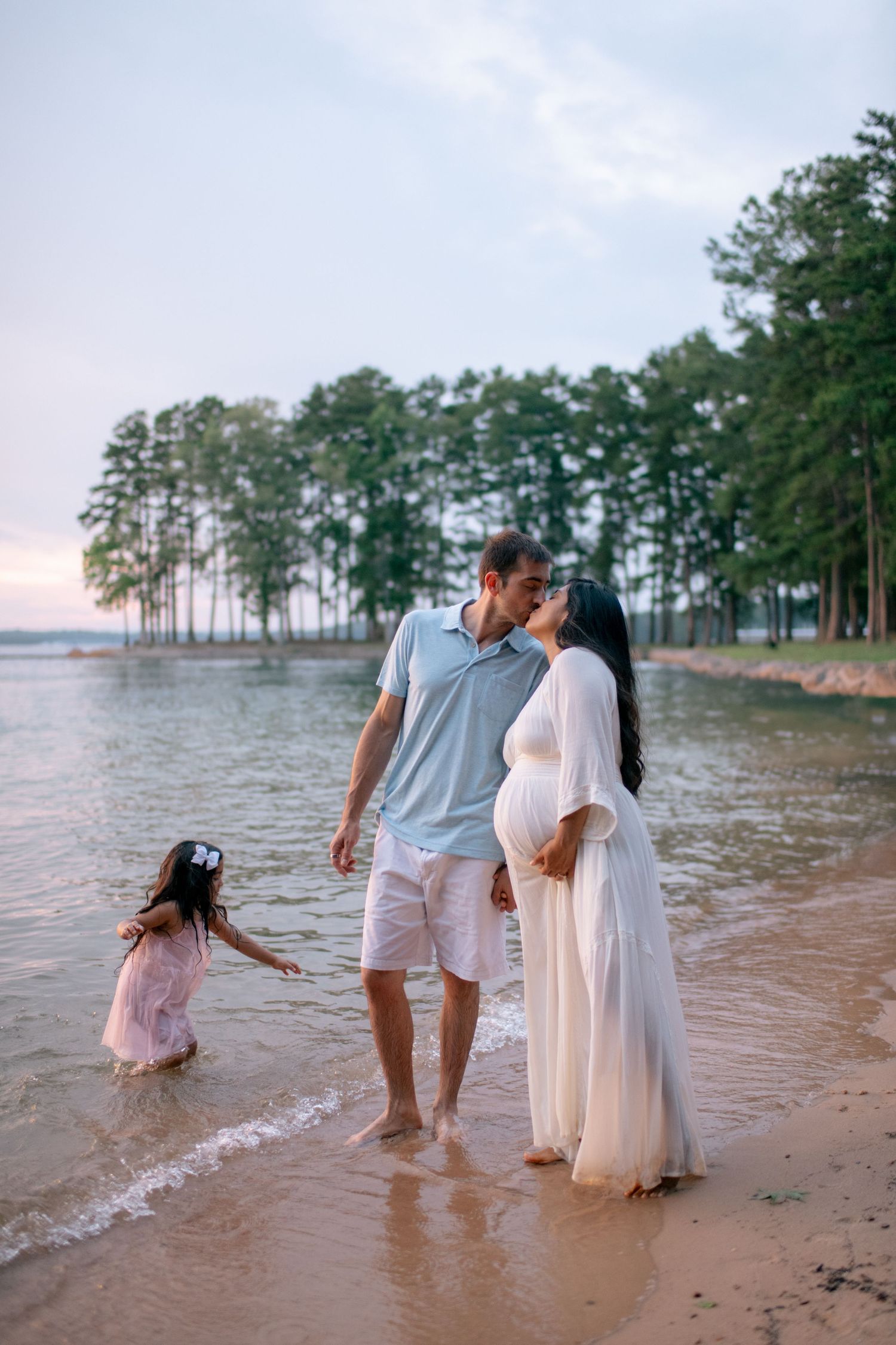 Family shares a tender moment while walking along a scenic lakeside beach at sunset with pine trees in the background.