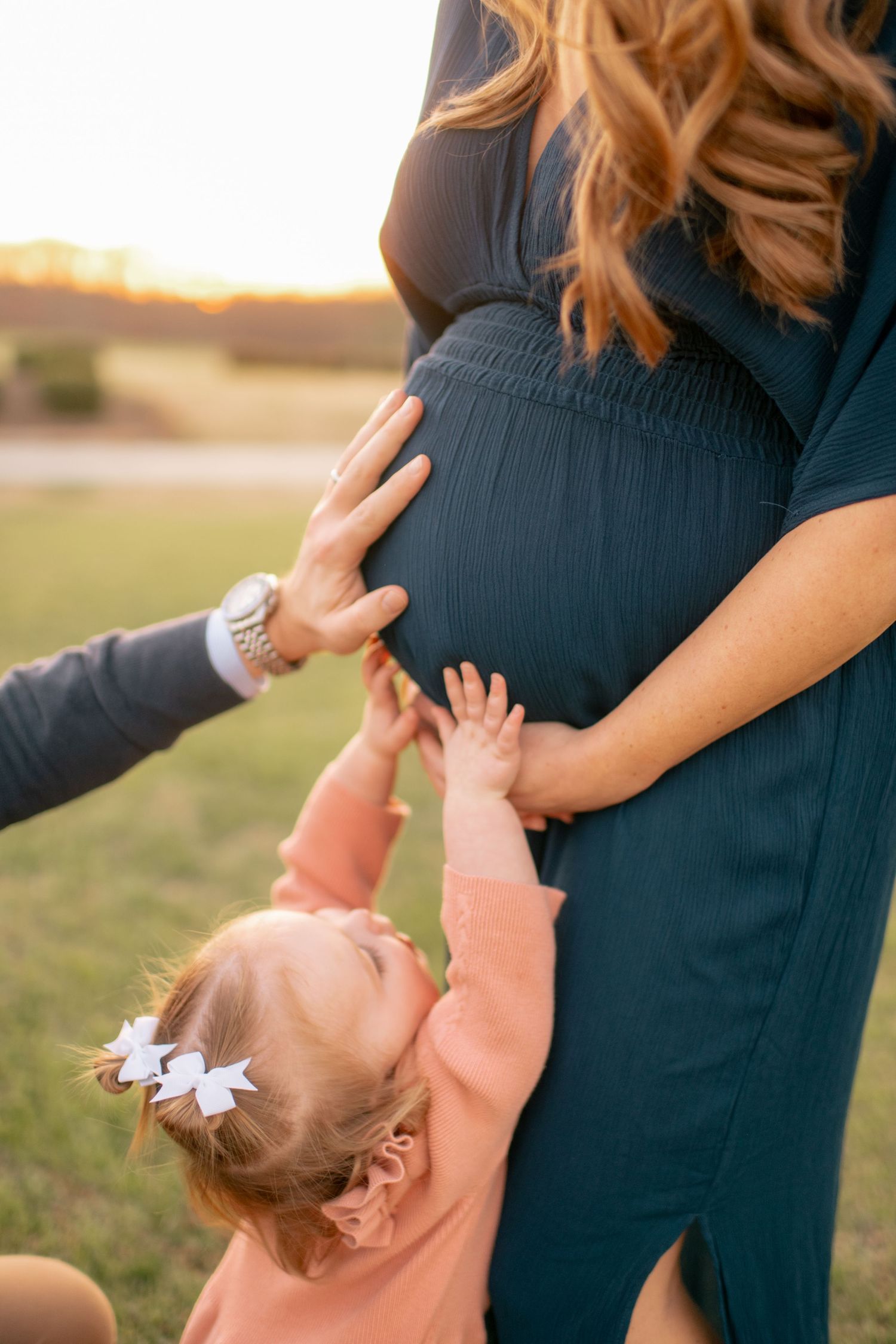 A toddler in pink with a hair bow reaches up to touch a navy blue maternity dress during golden hour sunset outdoors.