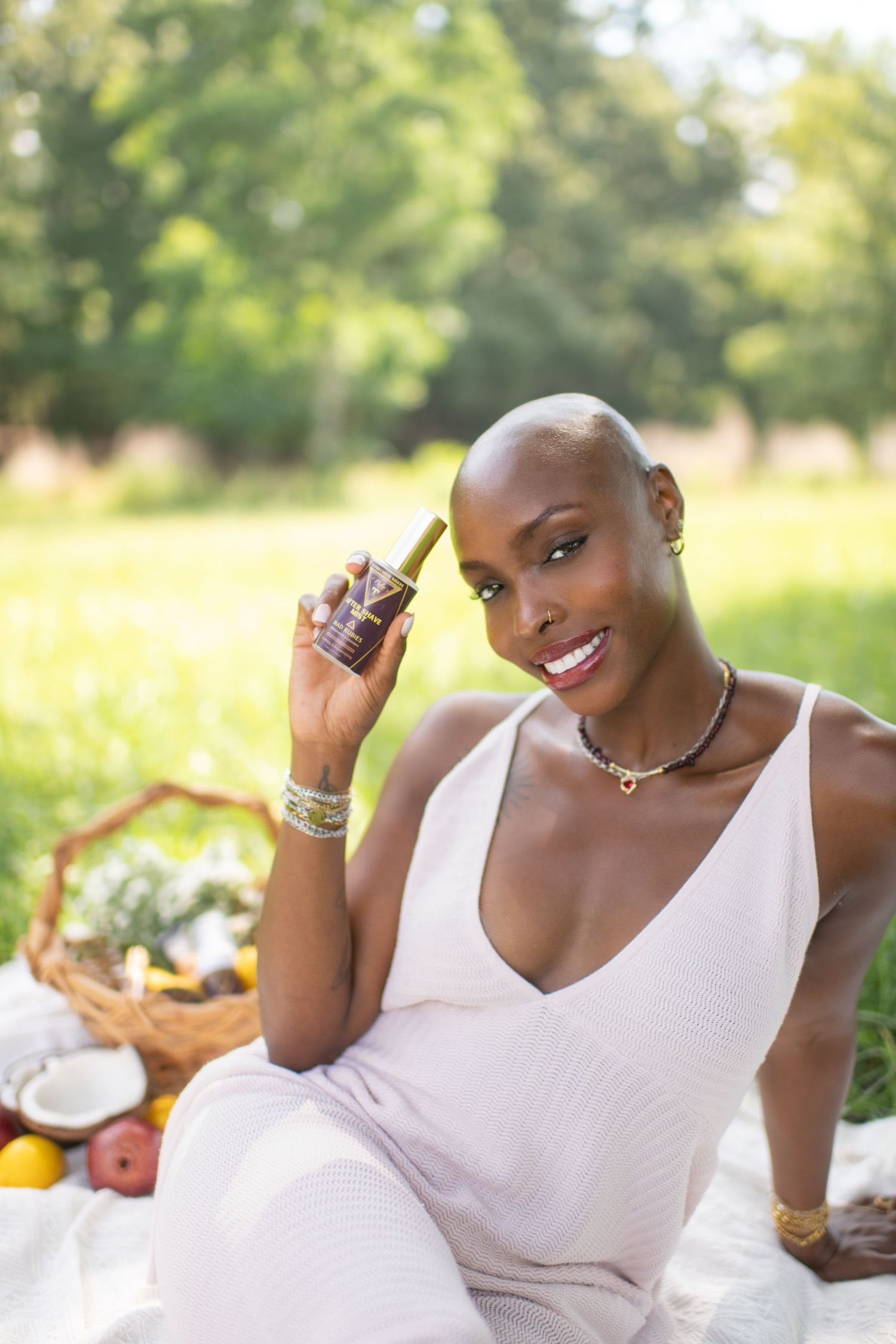 Smiling bald Black woman in white dress holding a serum bottle at a sunny outdoor picnic with fruits and a basket.