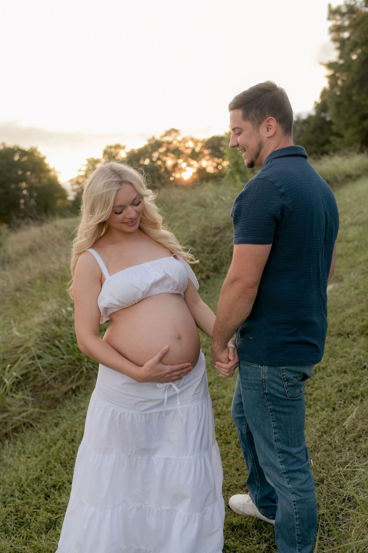 A couple poses together in a grassy field during sunset for a romantic maternity photoshoot.