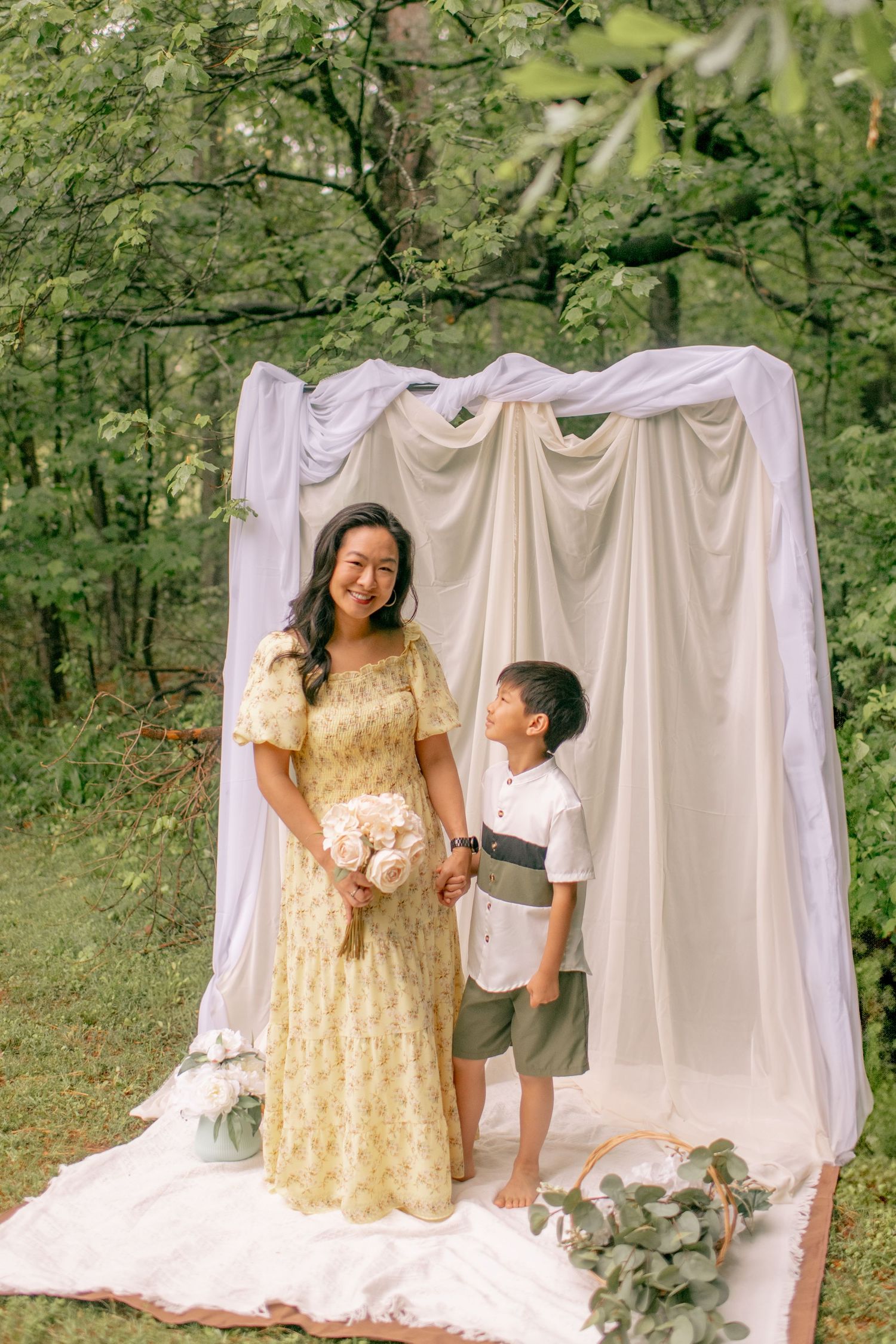 A yellow floral dress and white draped fabric backdrop create an elegant outdoor photo setting in a wooded area.