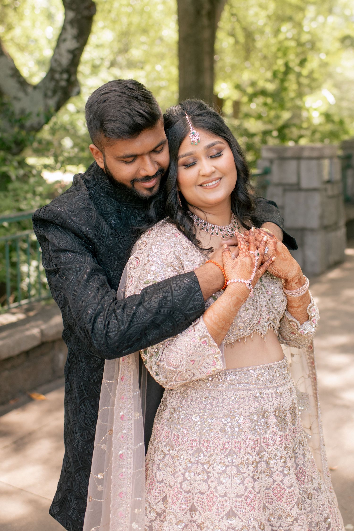 A romantic couple in traditional South Asian wedding attire embraces outdoors, wearing pink and black formal outfits.