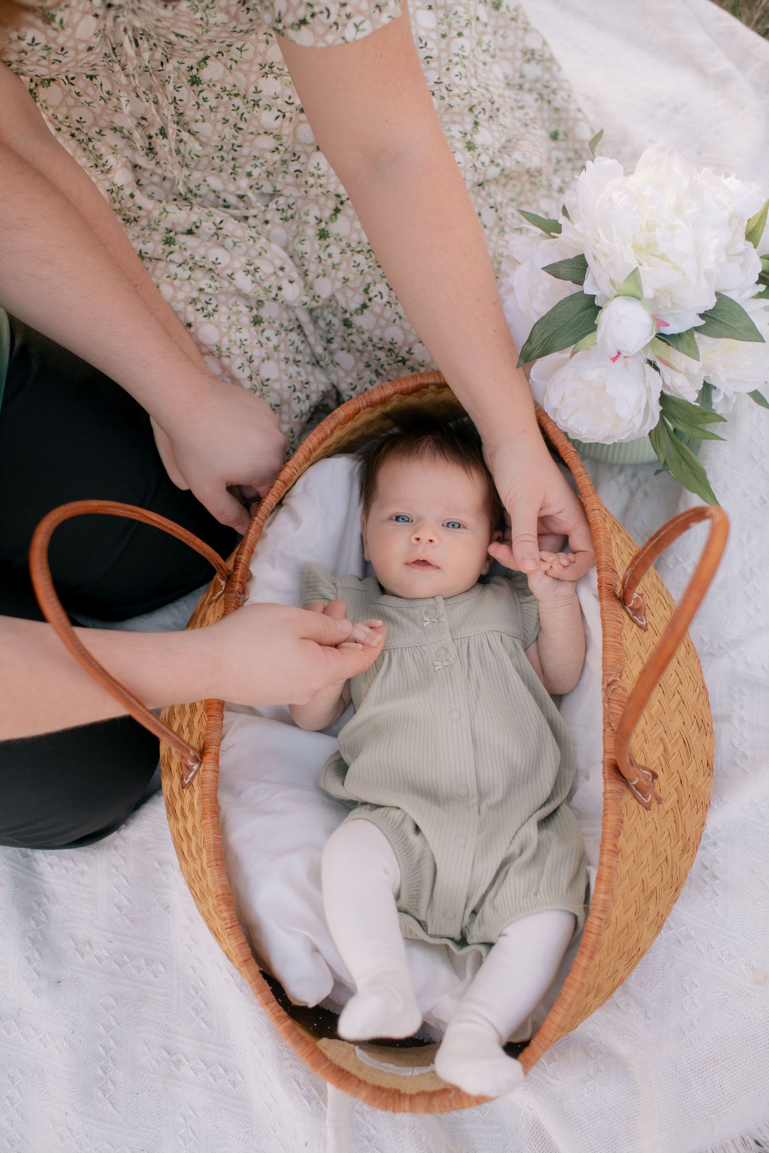 Baby wearing gray romper lying in woven brown basket on white blanket with hands reaching in and white flowers nearby.