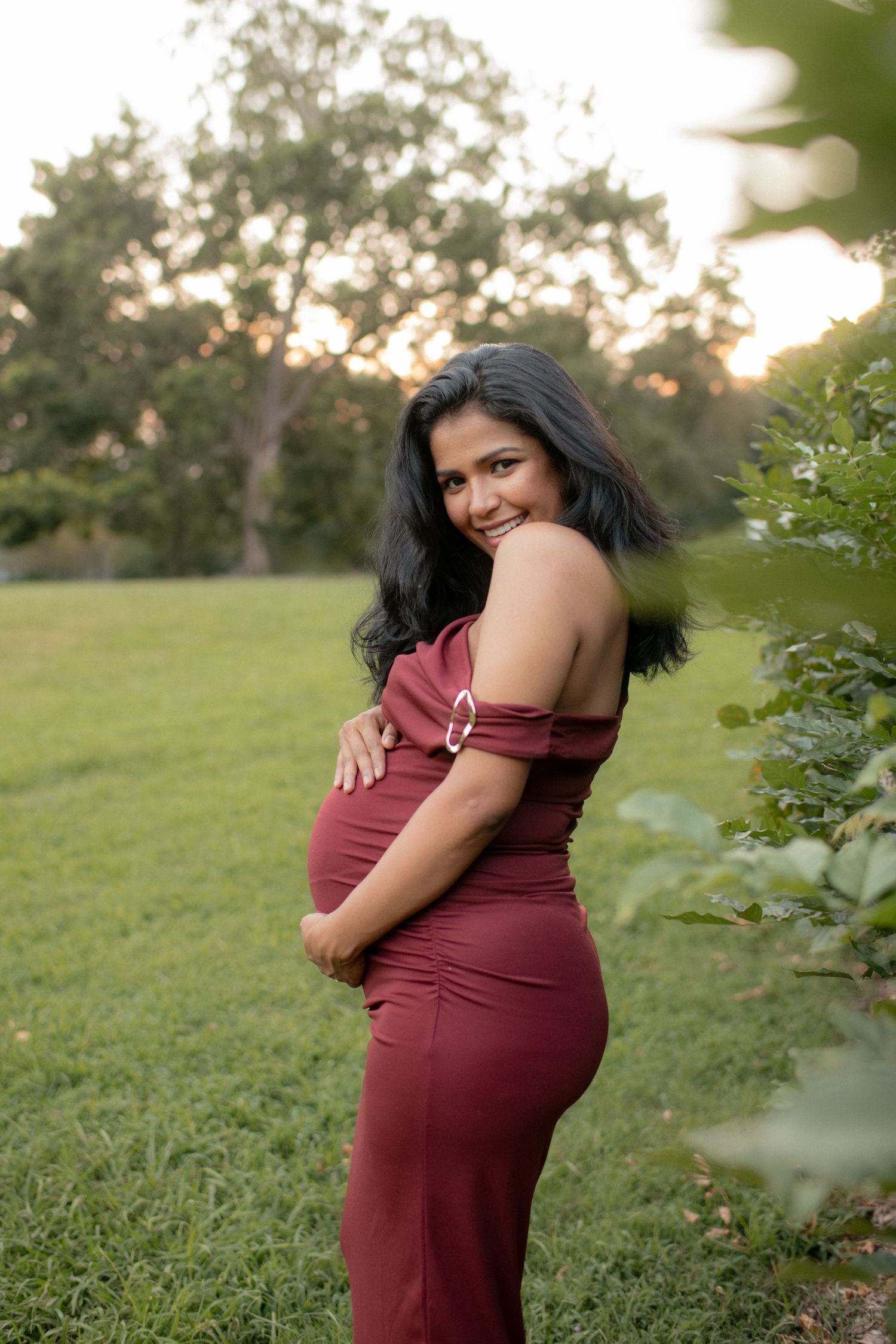 Side view of maternity poses in a burgundy dress against a natural outdoor setting.