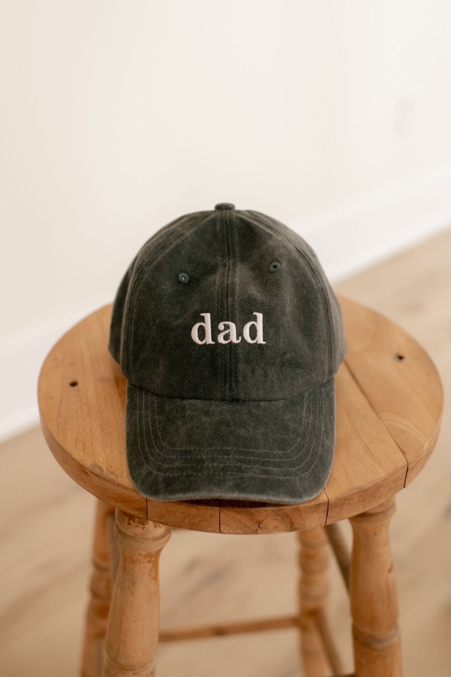 Charcoal gray baseball cap with 'dad' text sits on wooden stool.