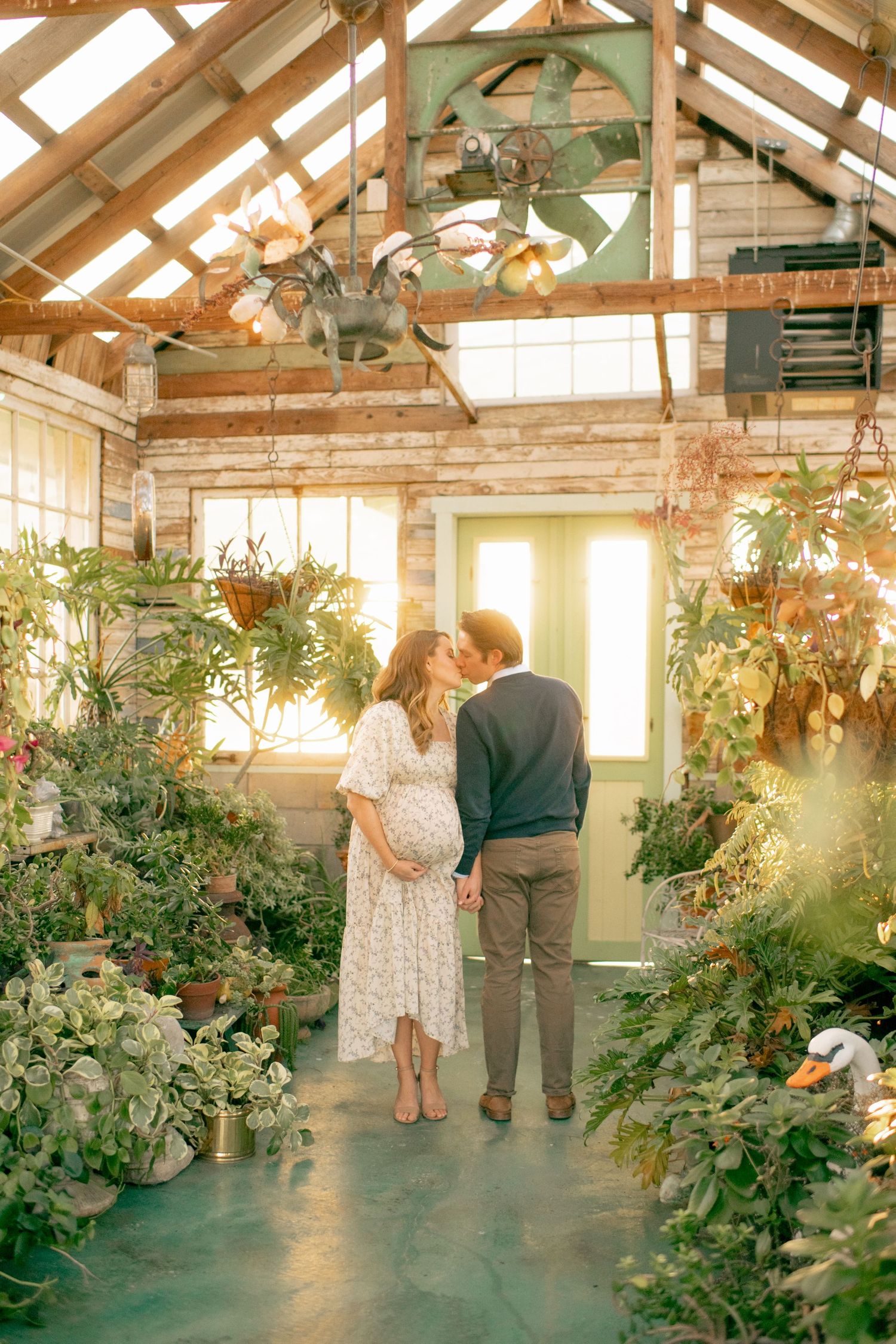A couple stands together in a sunlit greenhouse filled with lush plants and hanging baskets under wooden beams.