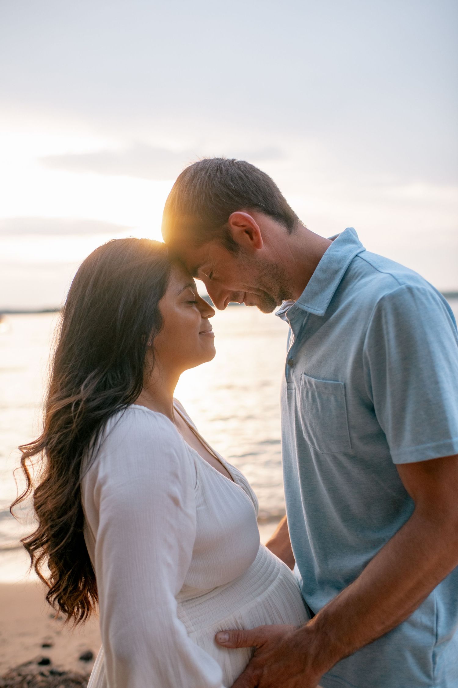 Romantic couple shares intimate moments at sunset on beach wearing blue shirt and white dress and embracing.