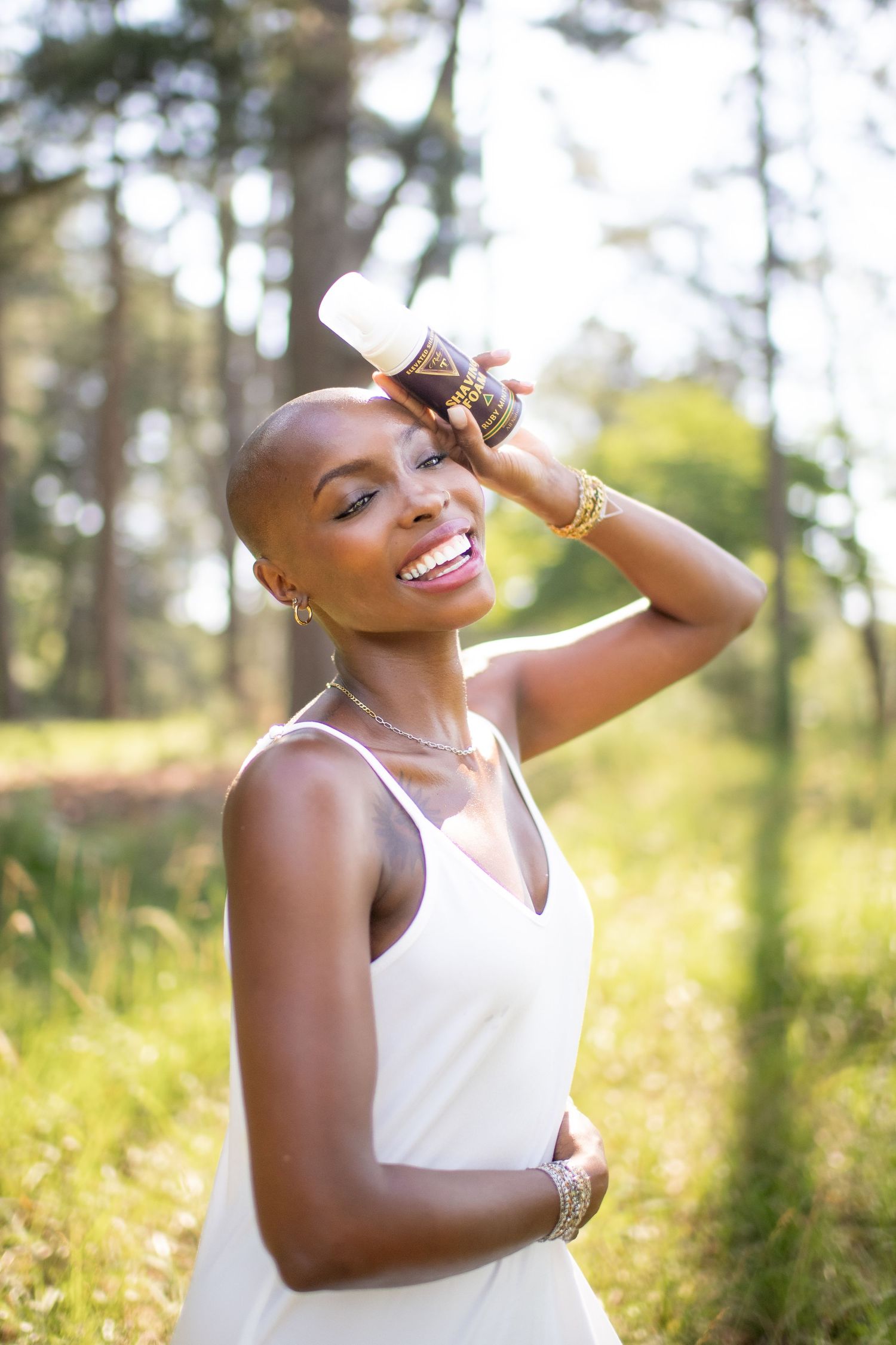 Smiling bald Black woman in white dress holding a bottle in a sunlit forest setting.