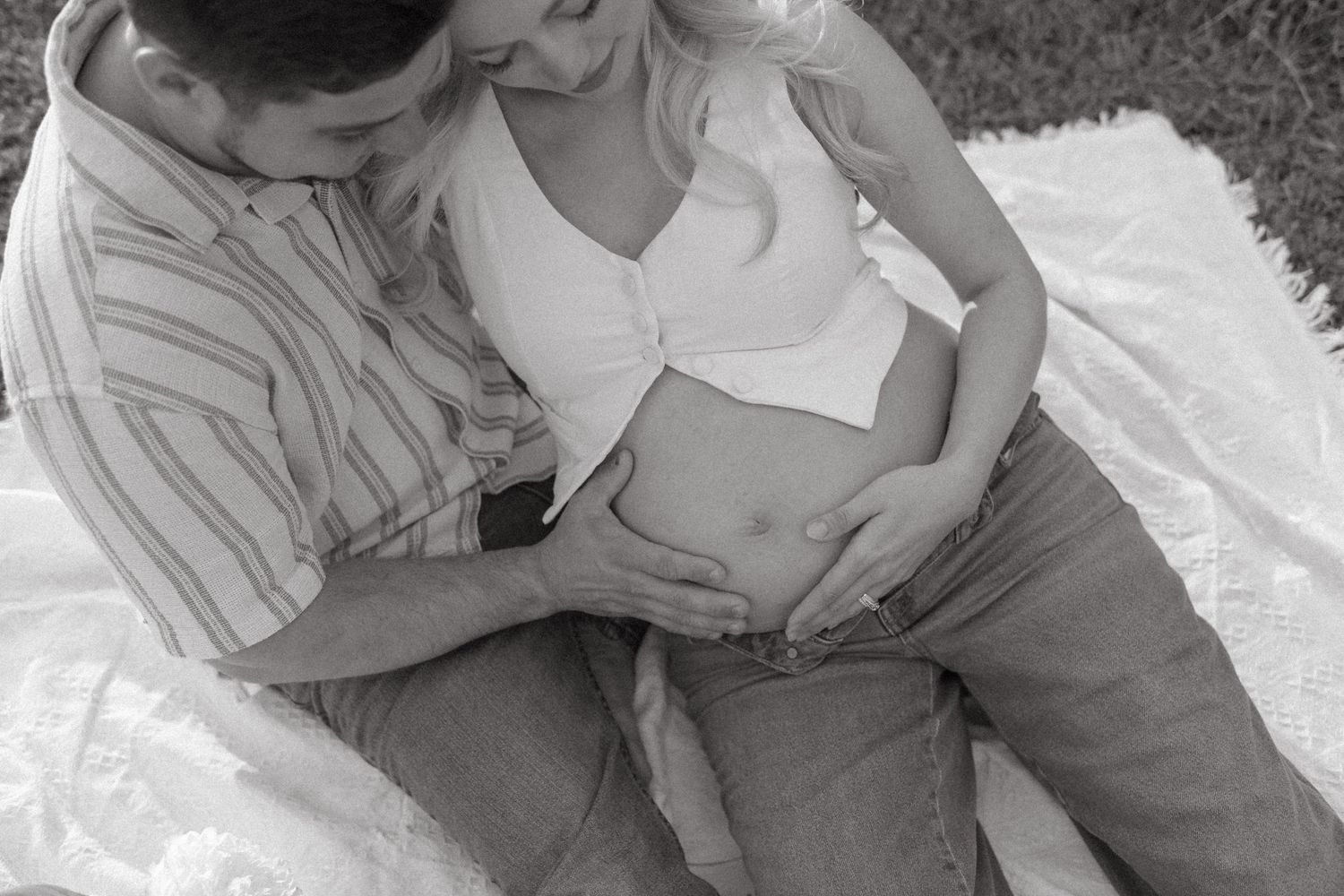 Maternity photoshoot on a grassy hillside at sunset with a white blanket laid out on the ground.
