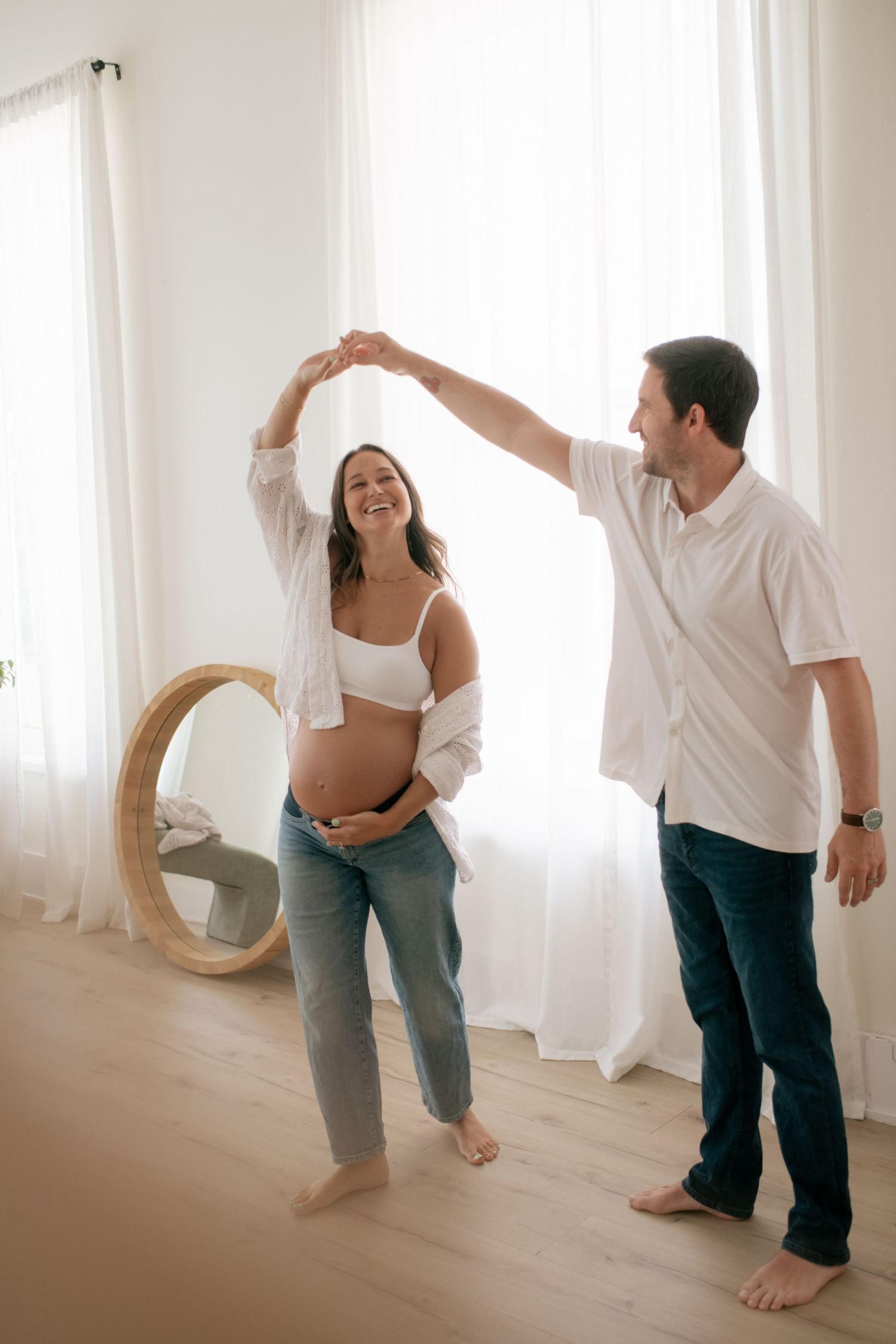 Playful maternity photoshoot with couple dancing and laughing by bright window.