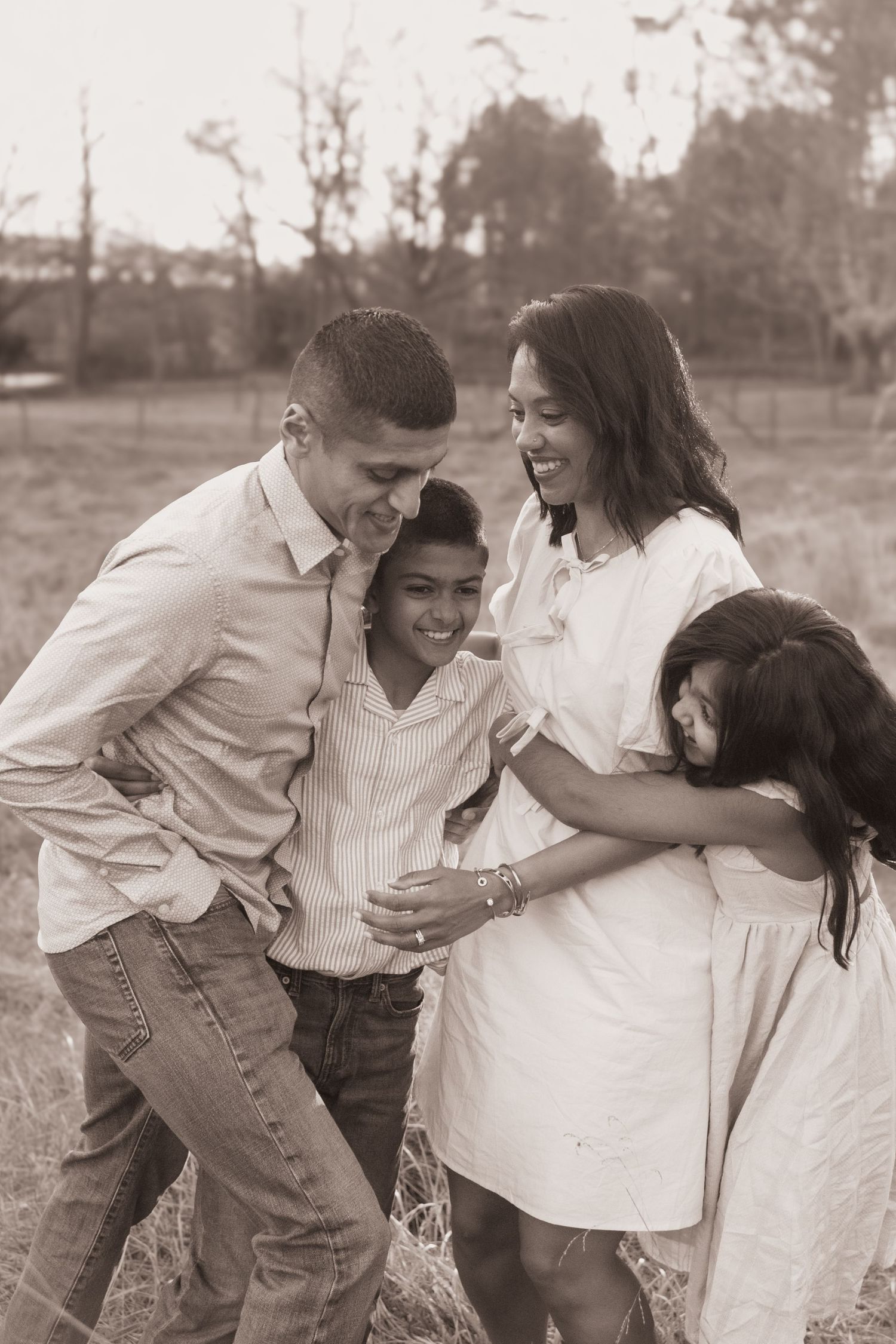A family in light-colored clothing shares a candid, joyful moment together outdoors in a sepia-toned photograph.