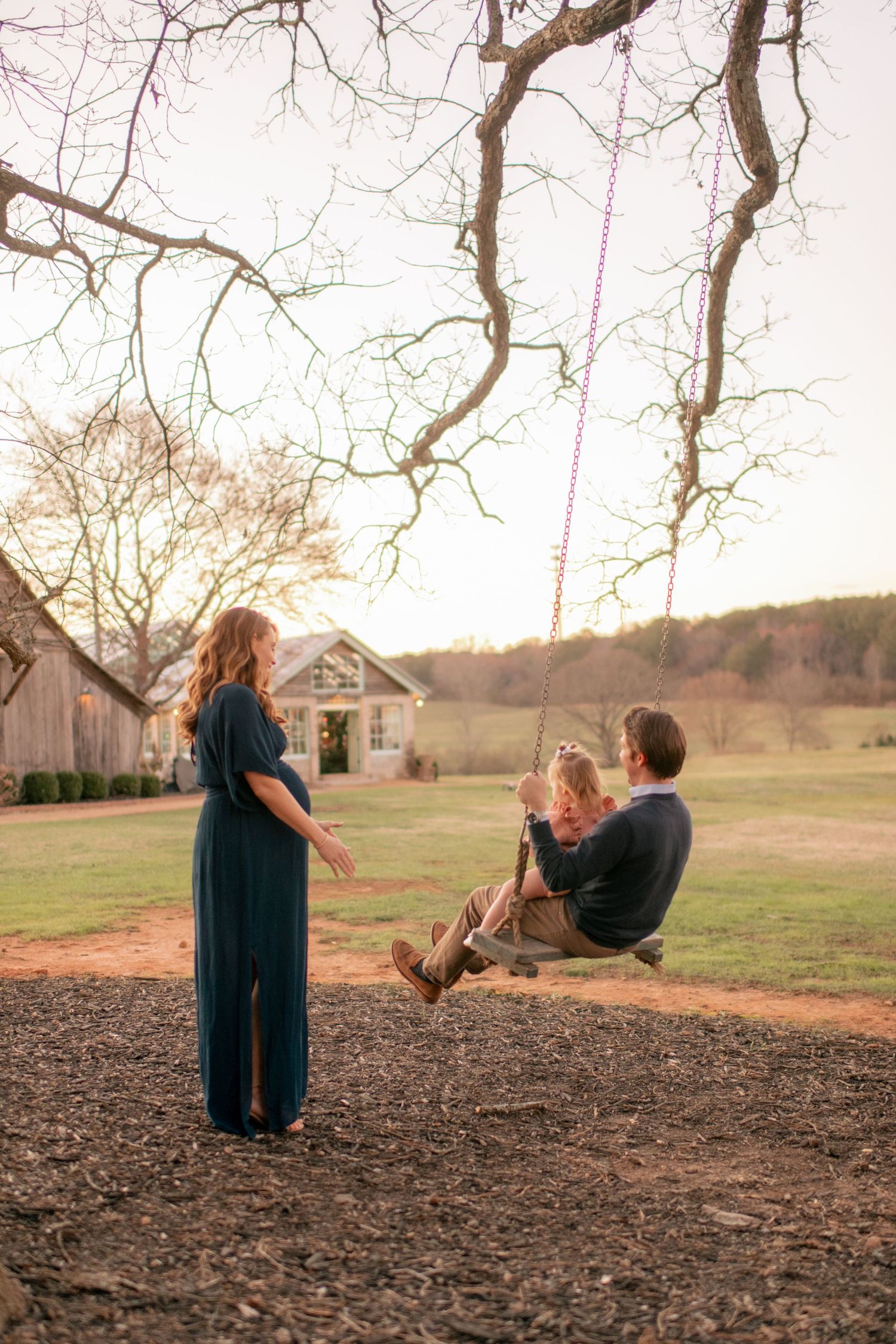 Family moment captured at sunset as figures play on tree swing in front of rustic farmhouse.