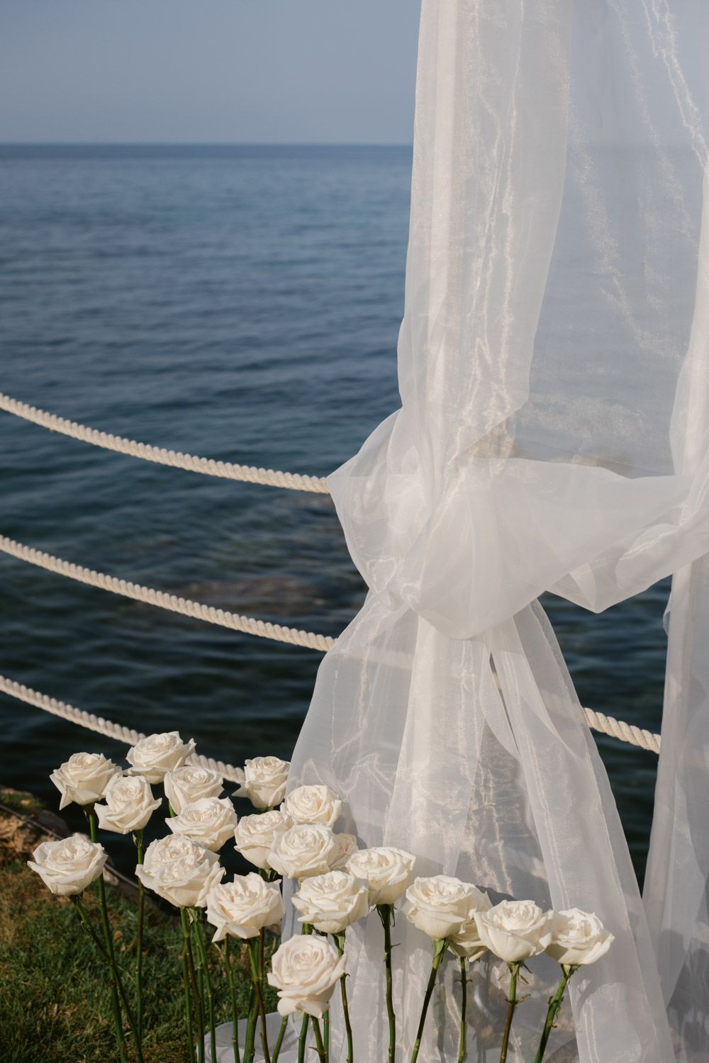 White sheer curtains and elegant white roses decorate an outdoor oceanfront wedding ceremony setup against blue waters.