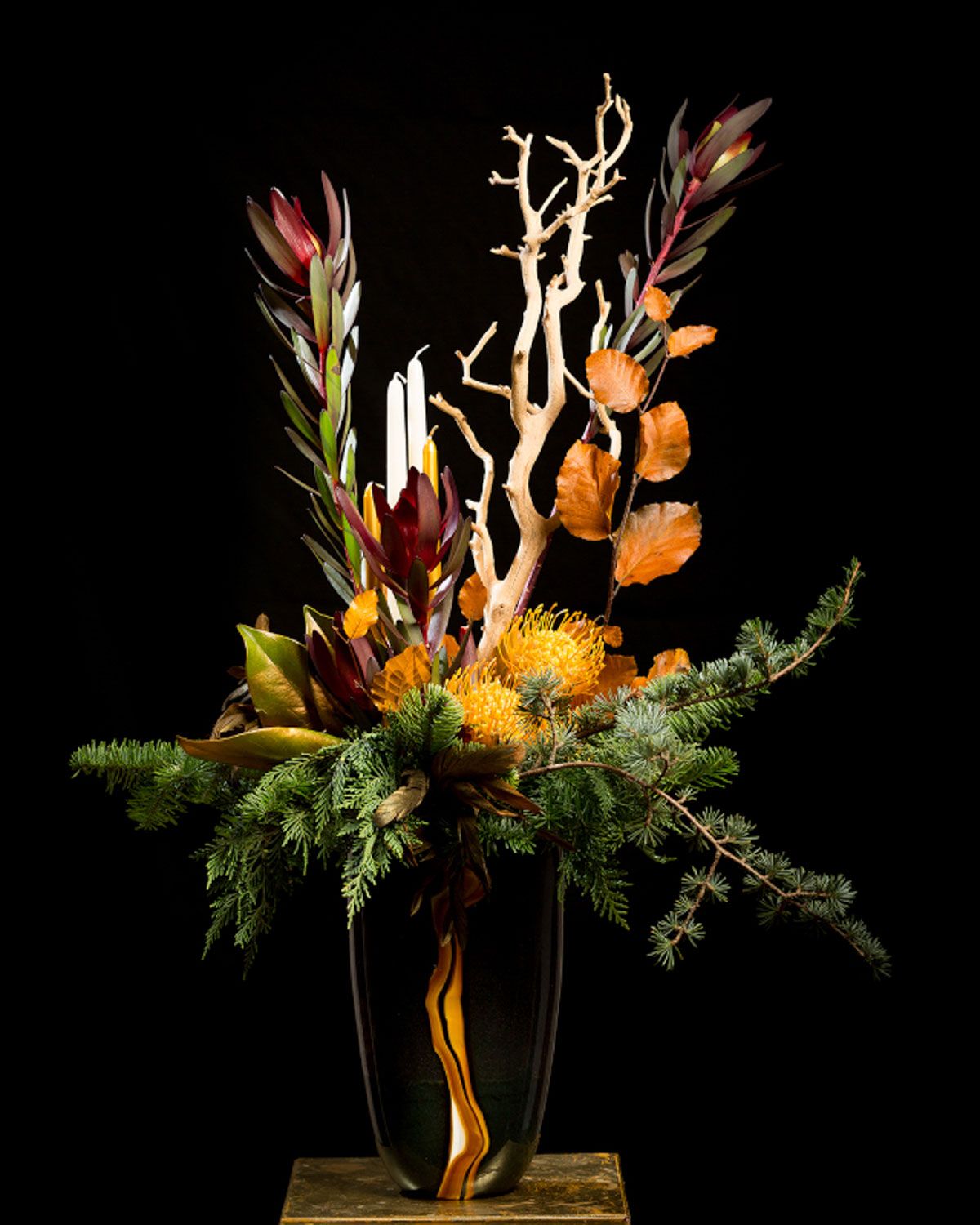 Modern floral arrangement with orange protea, white branches, and evergreen on wooden base.