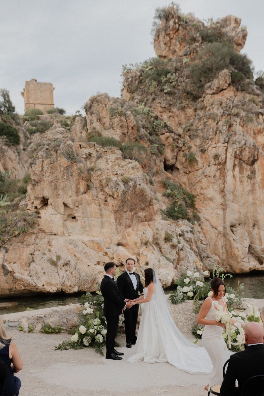A wedding ceremony takes place against dramatic rocky cliffs with elegant white floral arrangements and formal attire.