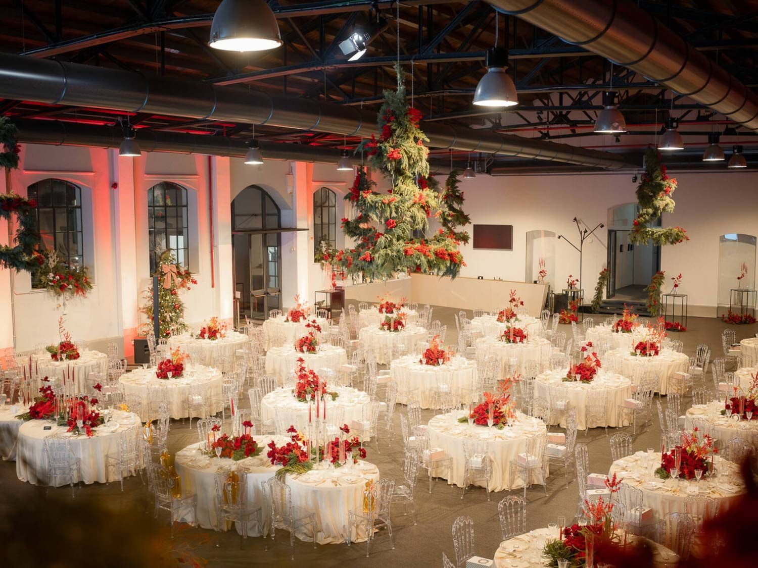 Corporate event flowers Italy. Large banquet hall setup with white tables and chairs decorated with red floral centerpieces.