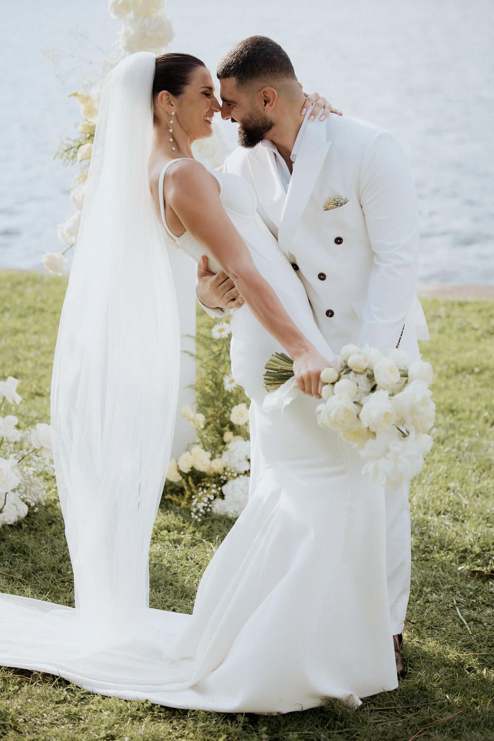 A romantic wedding couple in white attire embraces outdoors on a grassy lawn with a flowing veil and white bouquet.